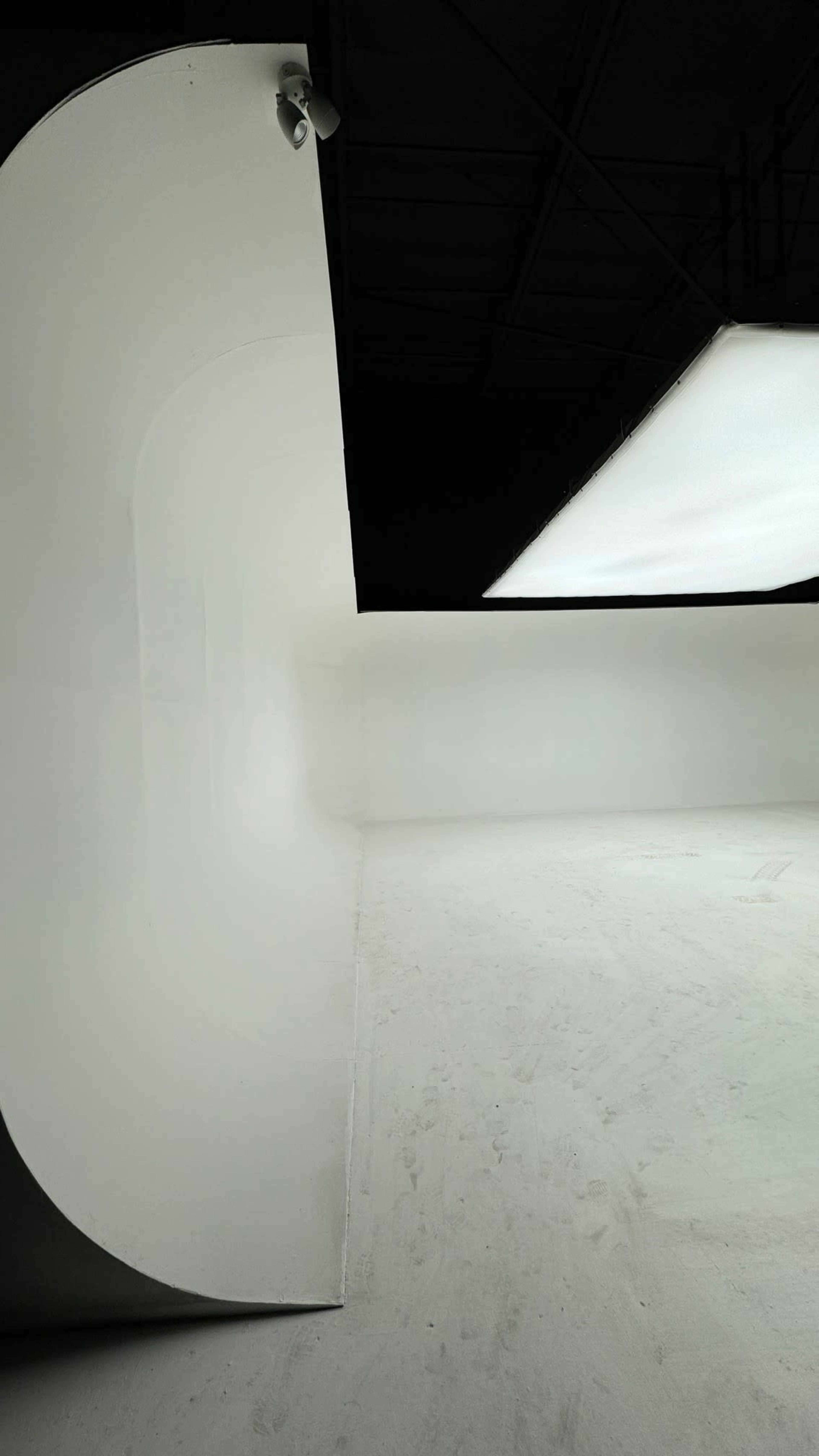 The image shows a minimalist interior space with curved white walls and a flat ceiling illuminated by a large light fixture.