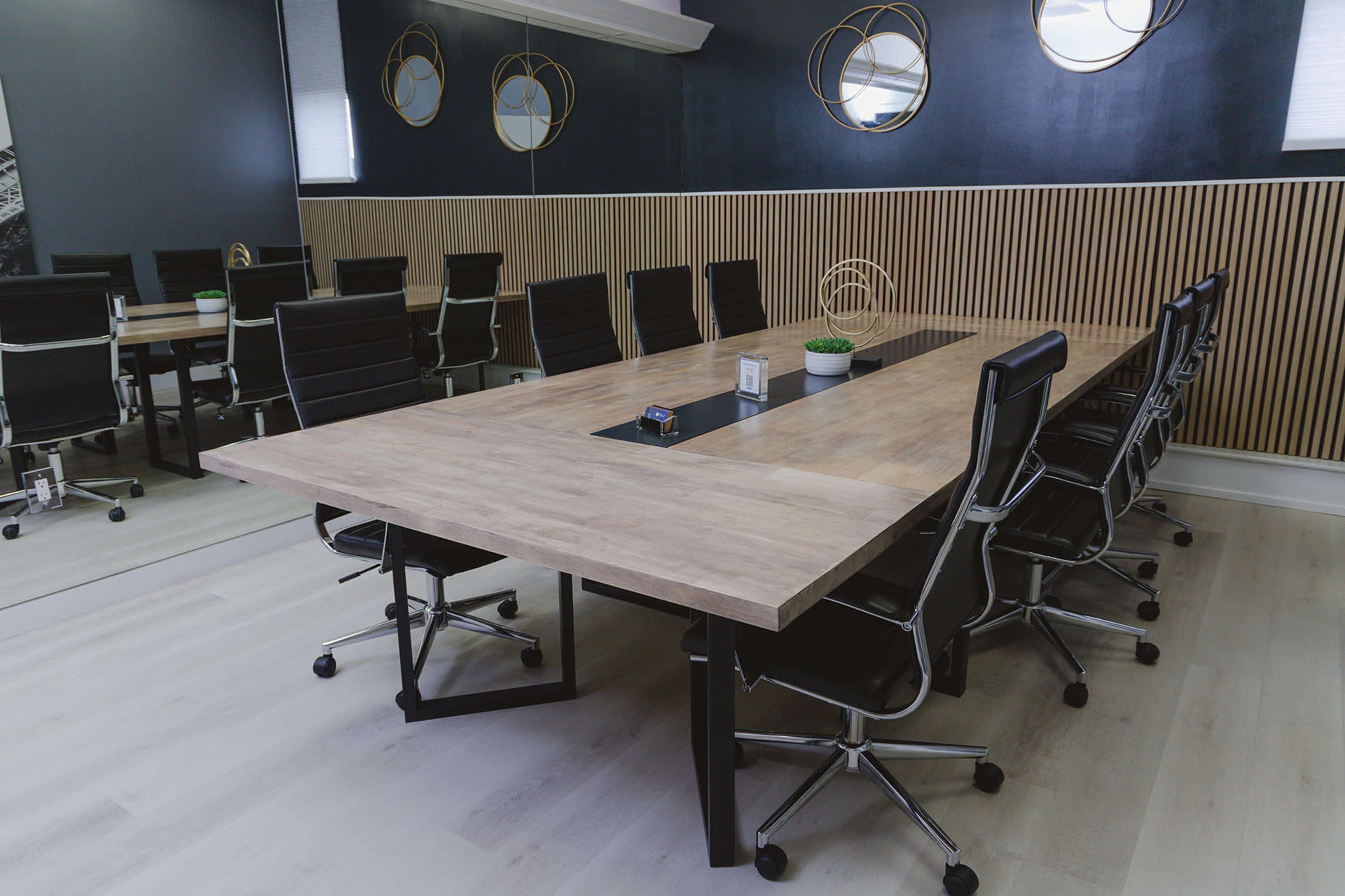 The image shows a modern conference room with a large wooden table surrounded by black rolling chairs and decorative mirrors on the wall.