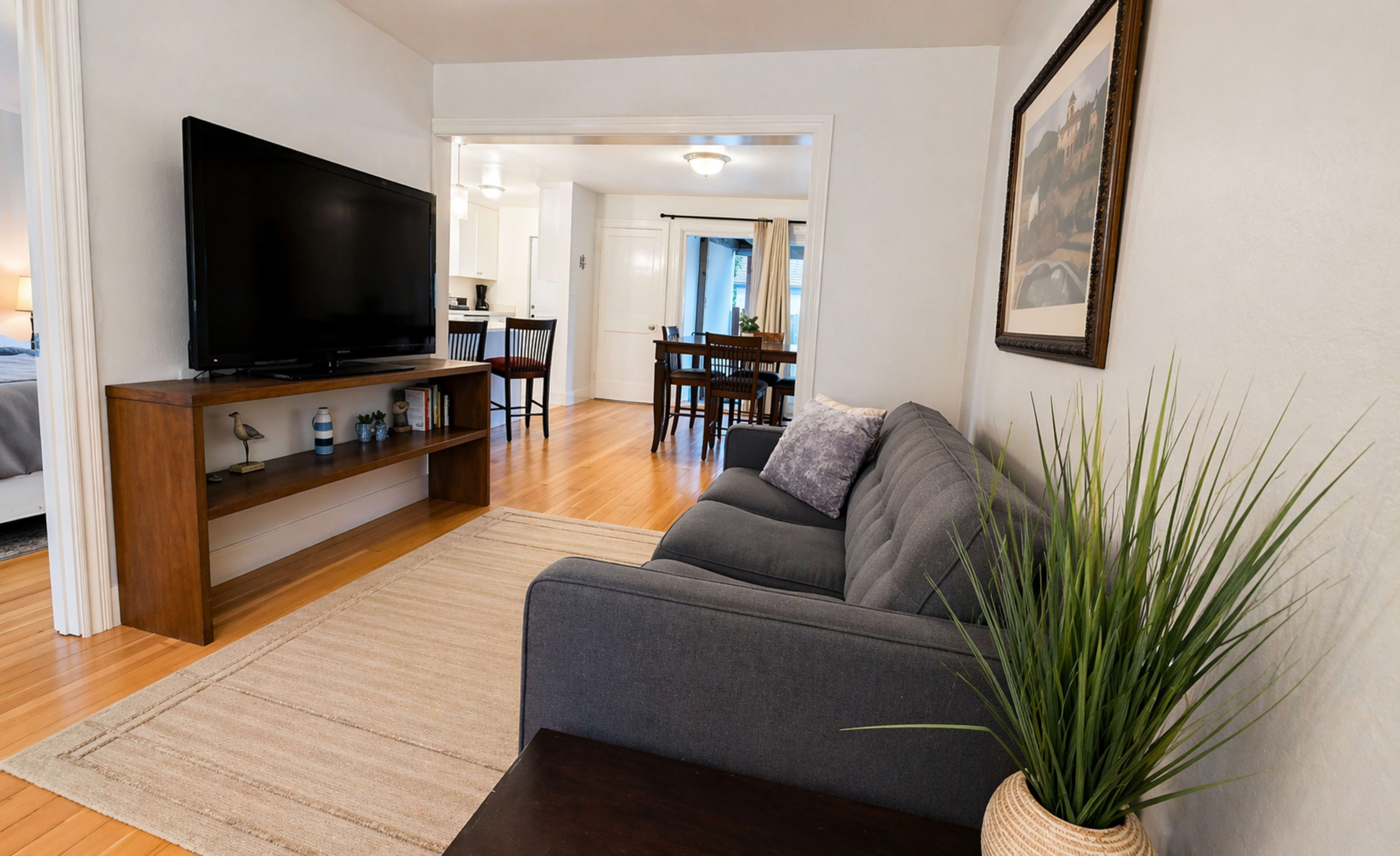 The image shows a cozy living room with a gray couch, a television on a wooden stand, and a plant in the corner, leading to a dining area visible through an adjacent doorway.