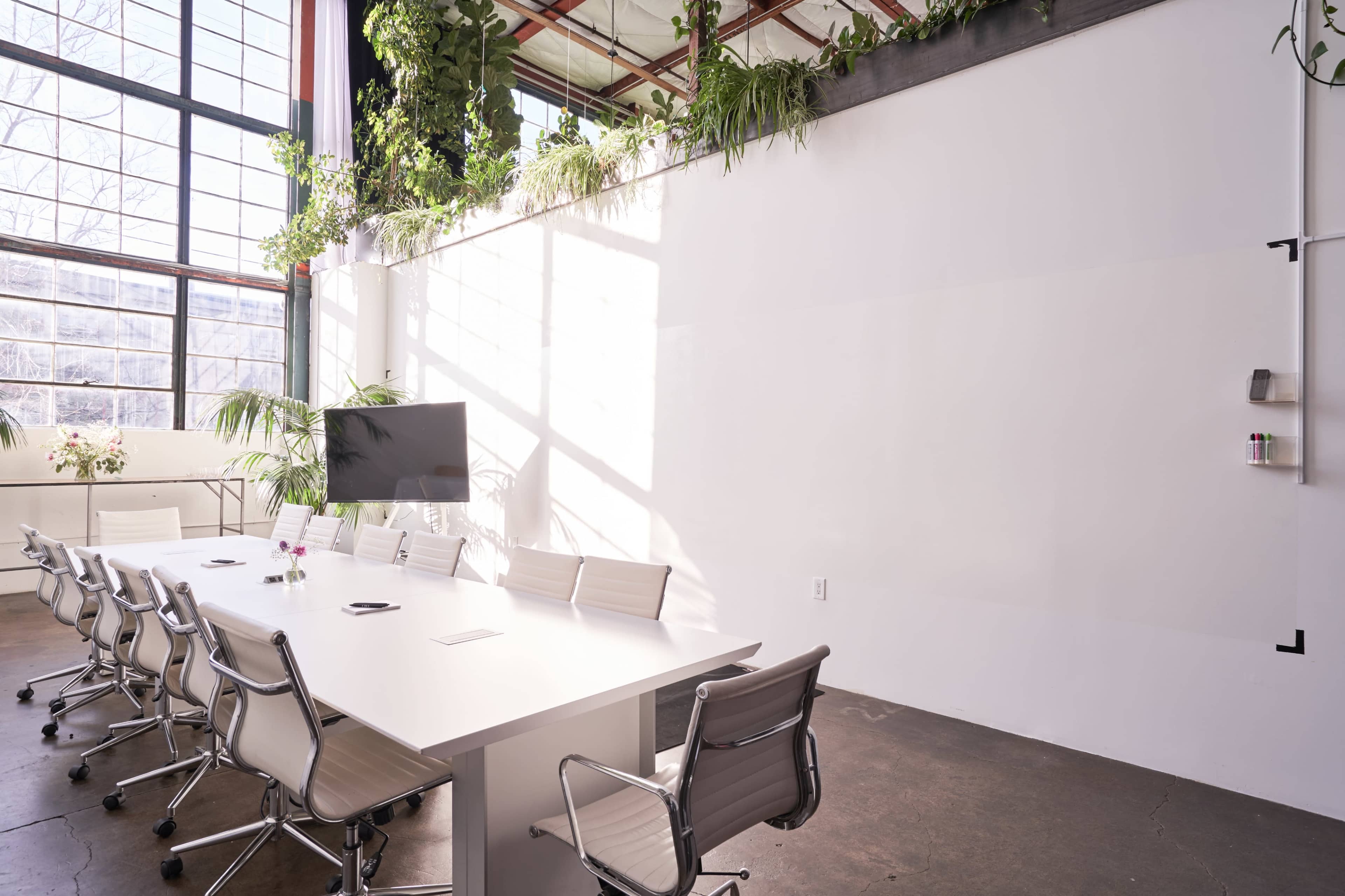 A bright conference room features a long white table surrounded by modern chairs, with large windows and plants lining the walls.