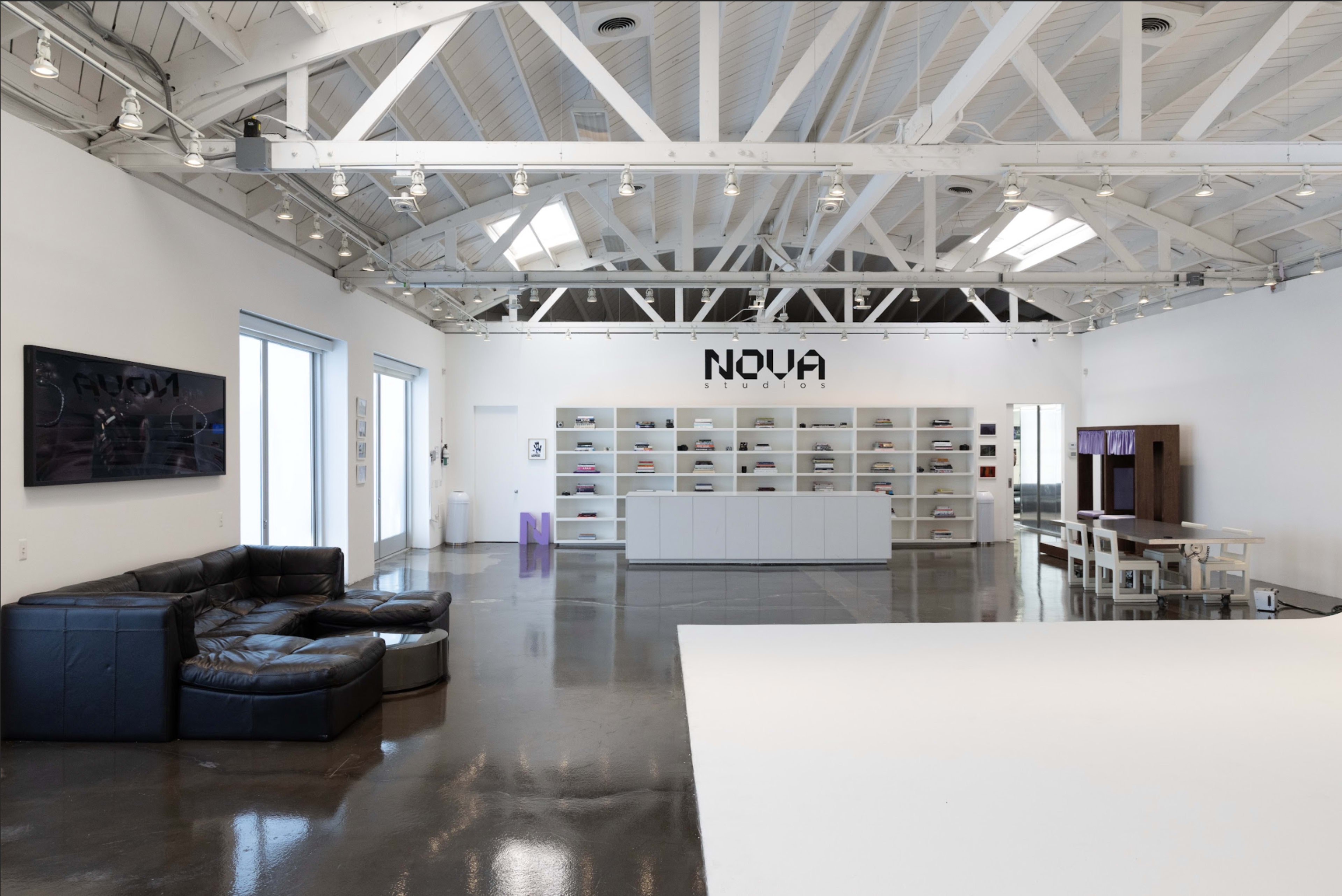 The image shows a spacious, modern studio with high ceilings, white walls, and a collection of shelves displaying various items in the background.