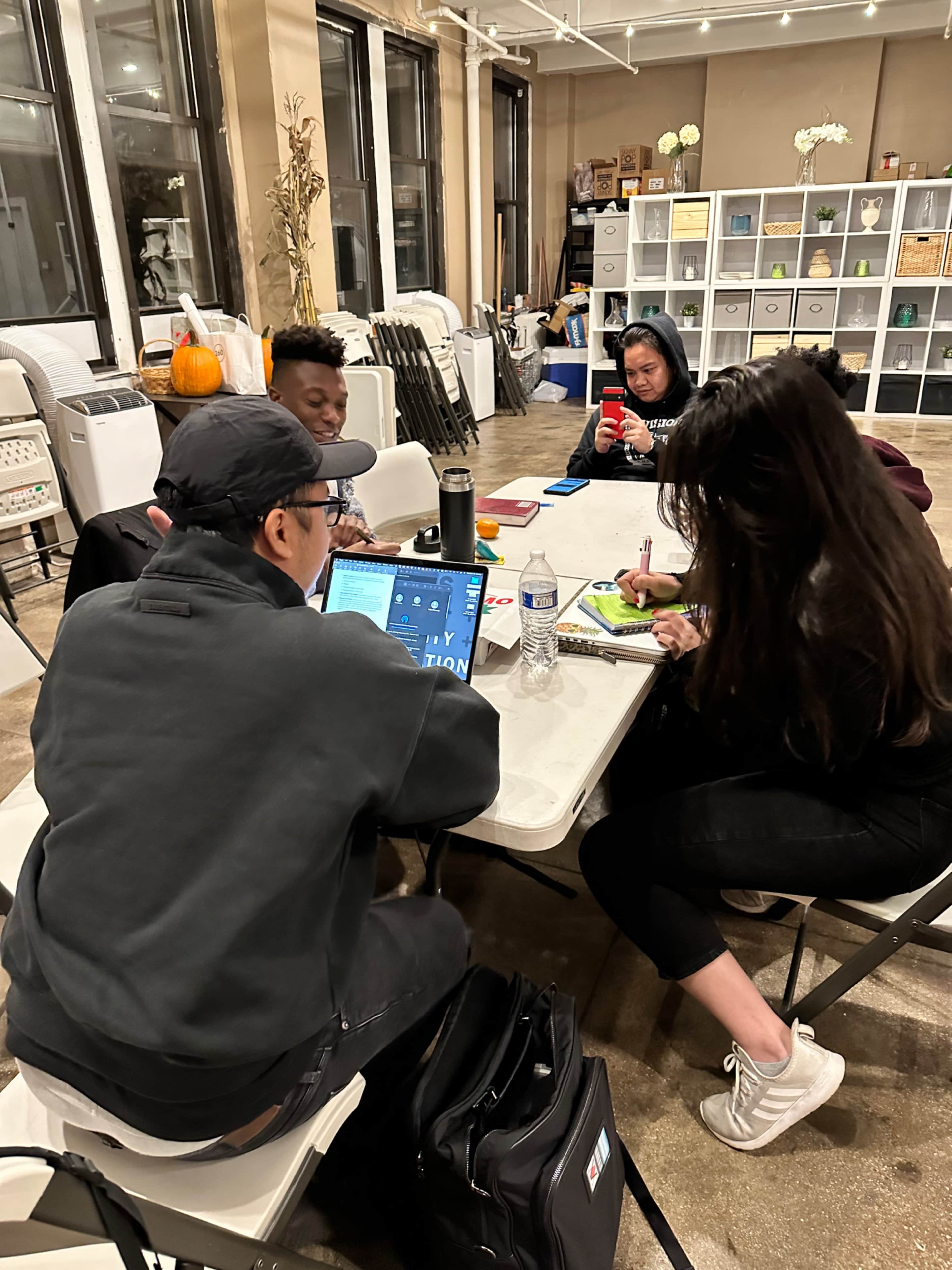 A group of four people collaborates around a table with laptops and notebooks in a well-lit indoor space.