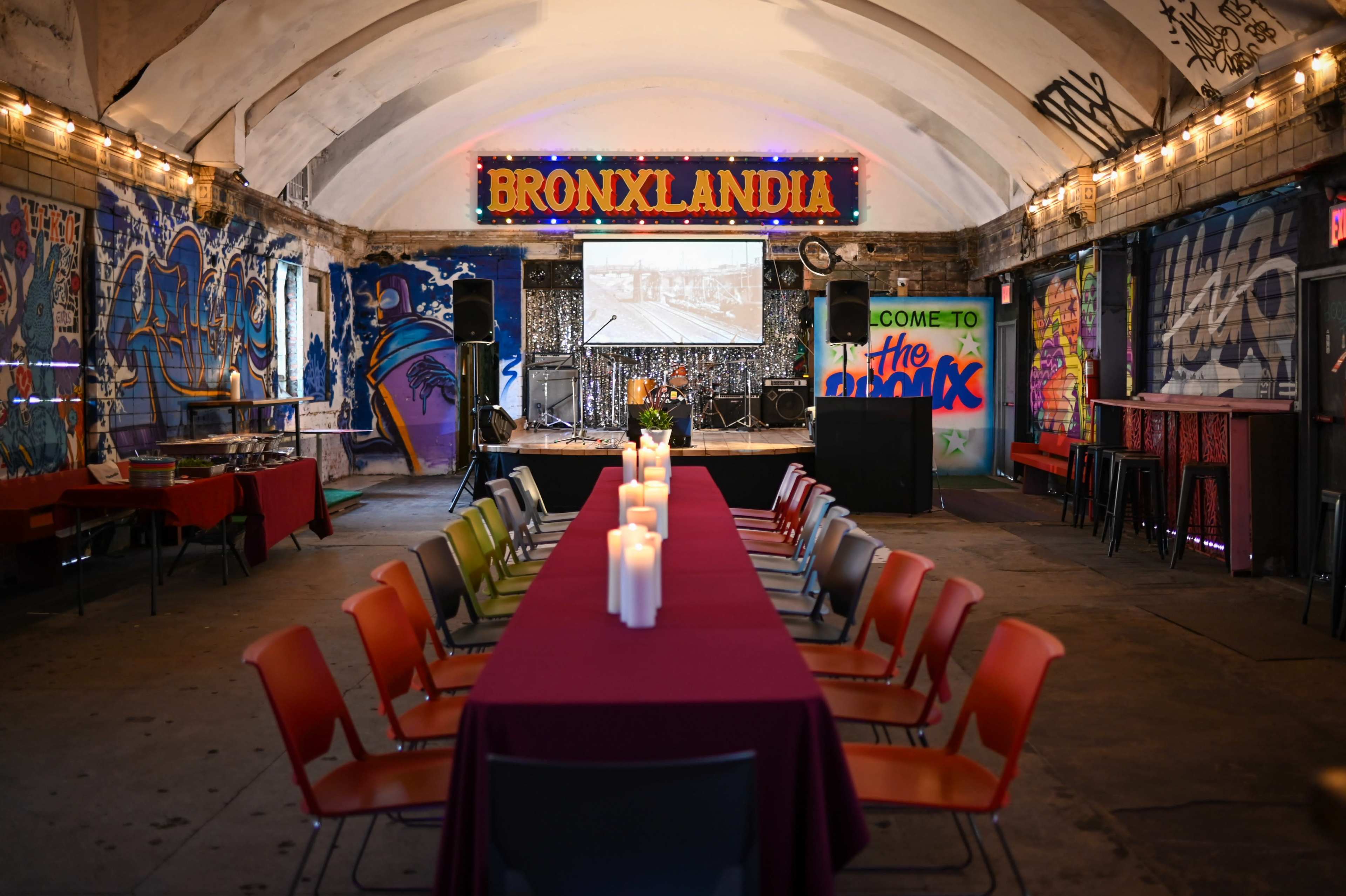 The image shows an indoor event space called "Bronxlandia," featuring a long table set with candles and colorful chairs, surrounded by vibrant graffiti and a stage with musical equipment.