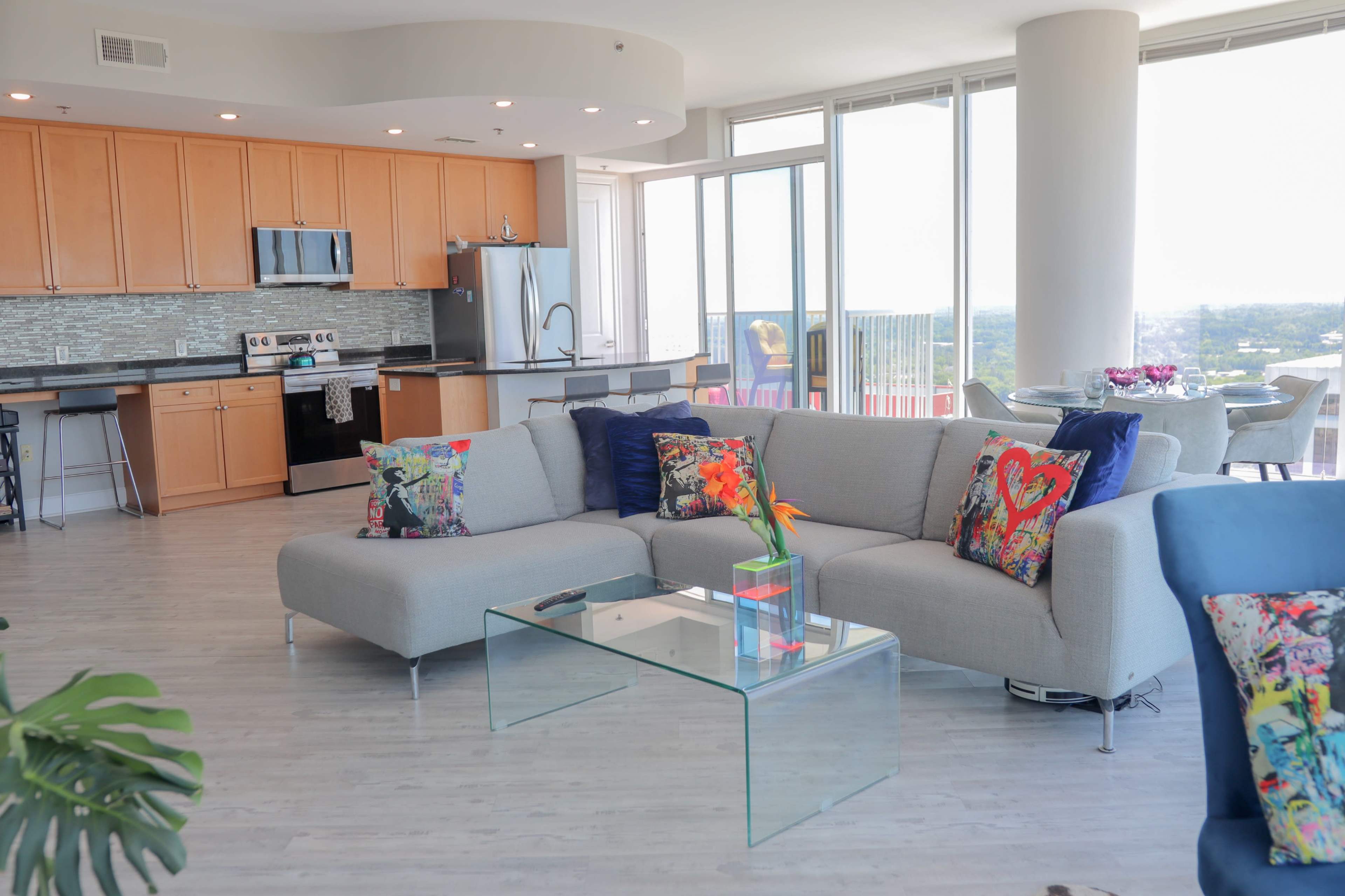 A modern living room with a grey sectional sofa, colorful throw pillows, a glass coffee table, and large windows offering a view of the skyline.