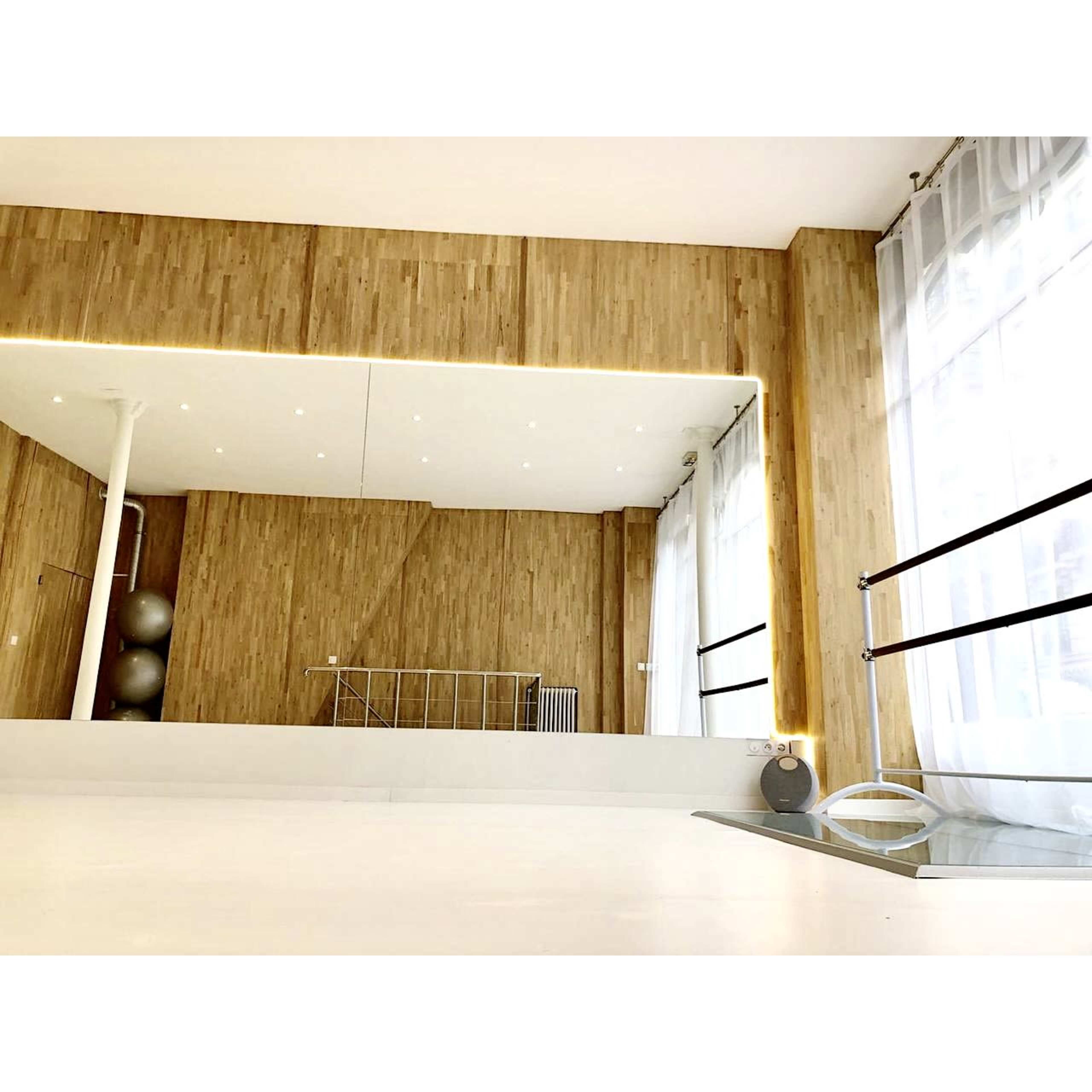 A bright, modern room with wooden walls, a large mirror, and floor-to-ceiling windows, featuring minimalistic ballet barres.