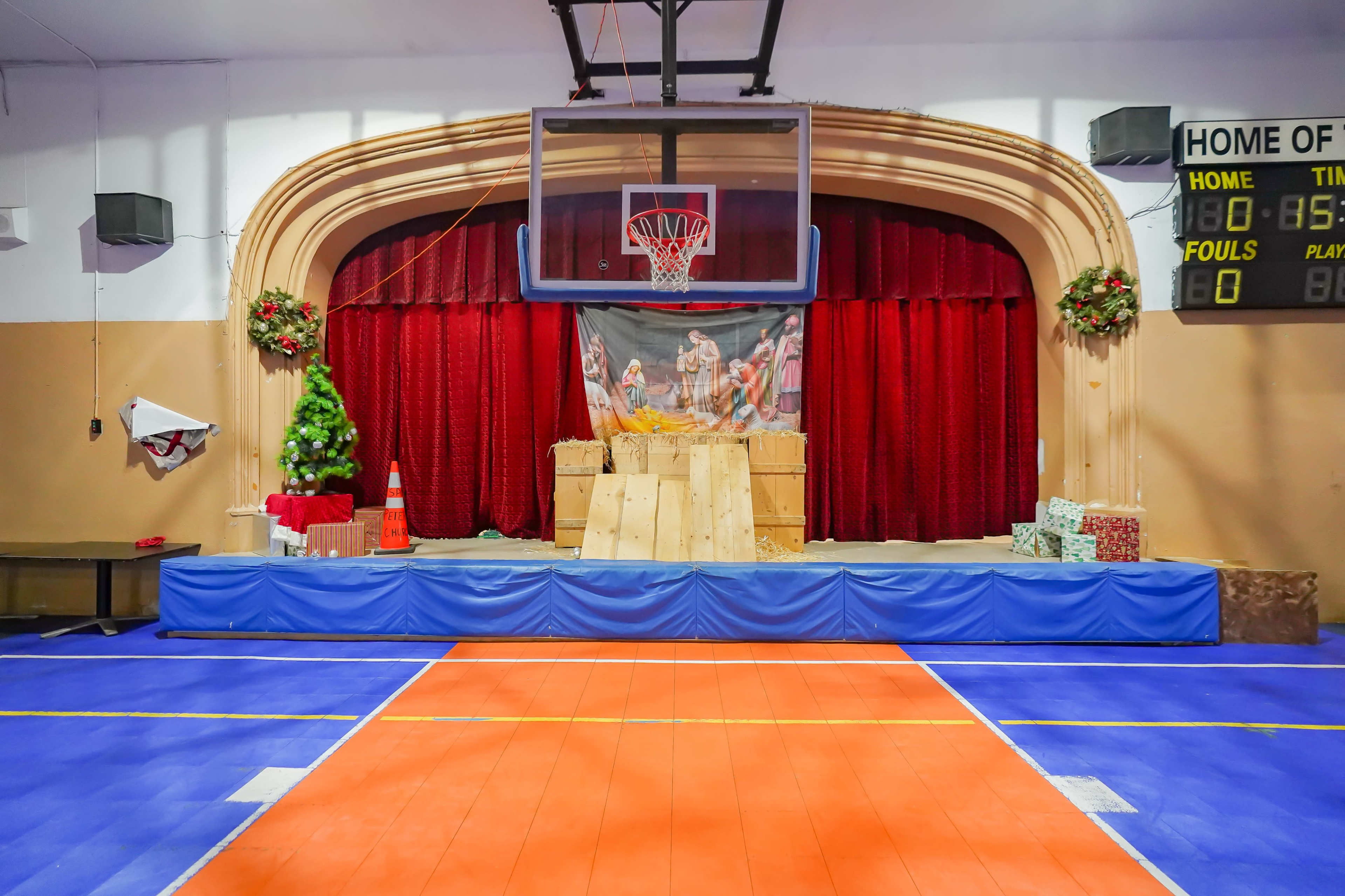 A basketball hoop is positioned above a decorated stage with a backdrop featuring a holiday scene, surrounded by wooden crates and gift boxes.