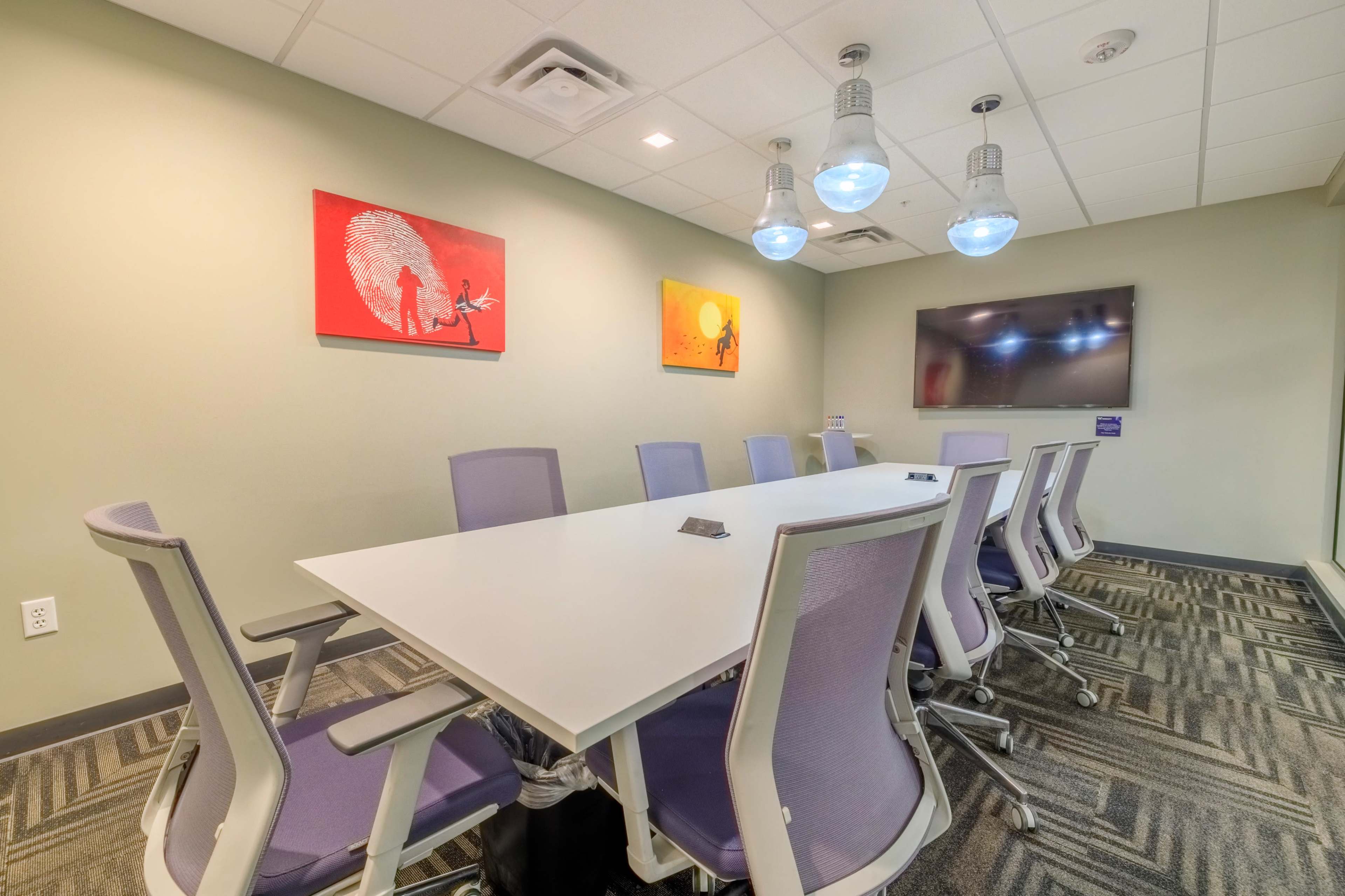 The image shows a modern conference room featuring a large white table surrounded by purple chairs, colorful wall art, and a flat-screen monitor.
