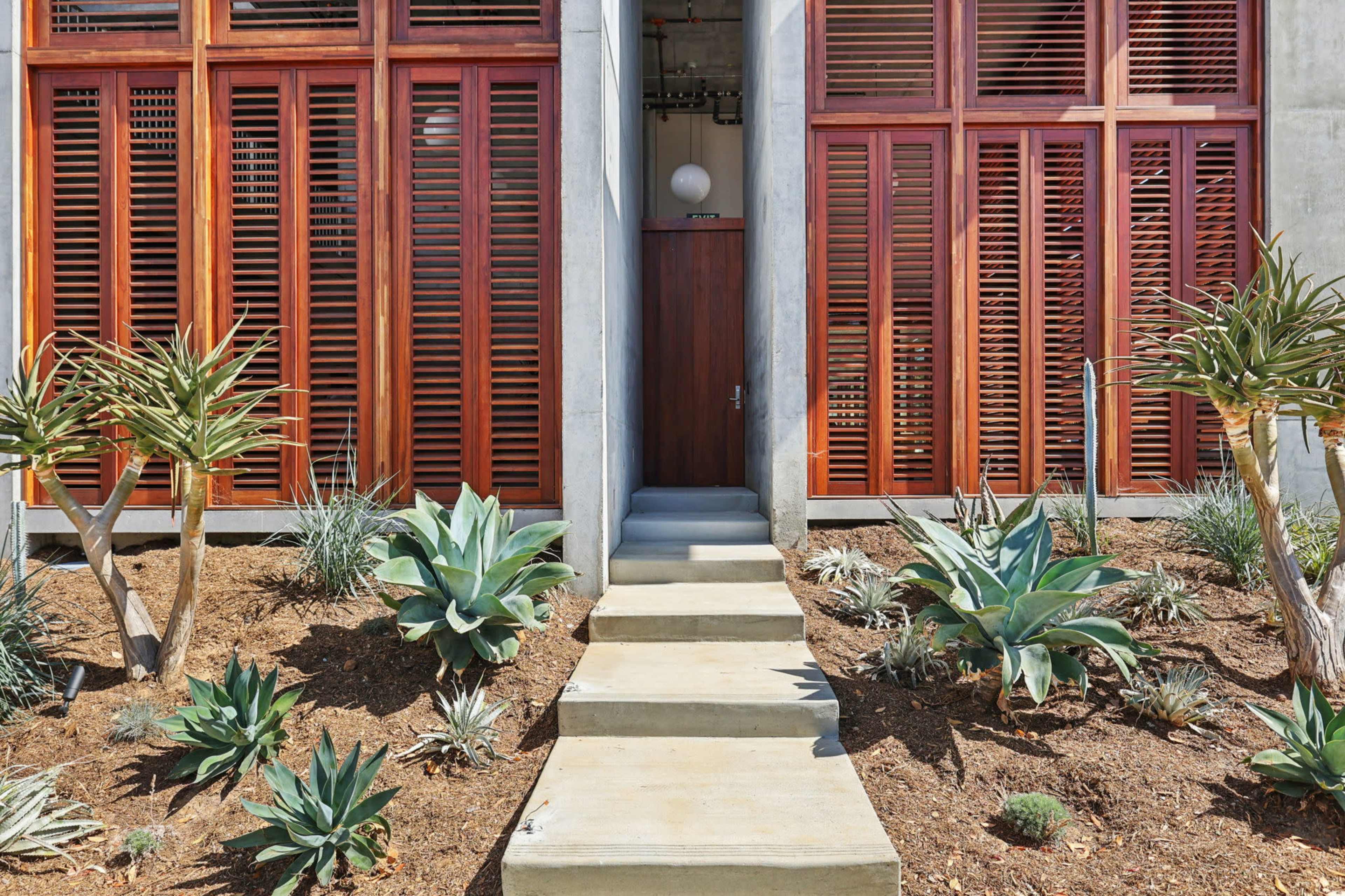A pathway leads to a door framed by wooden shutters and surrounded by various desert plants and succulents.