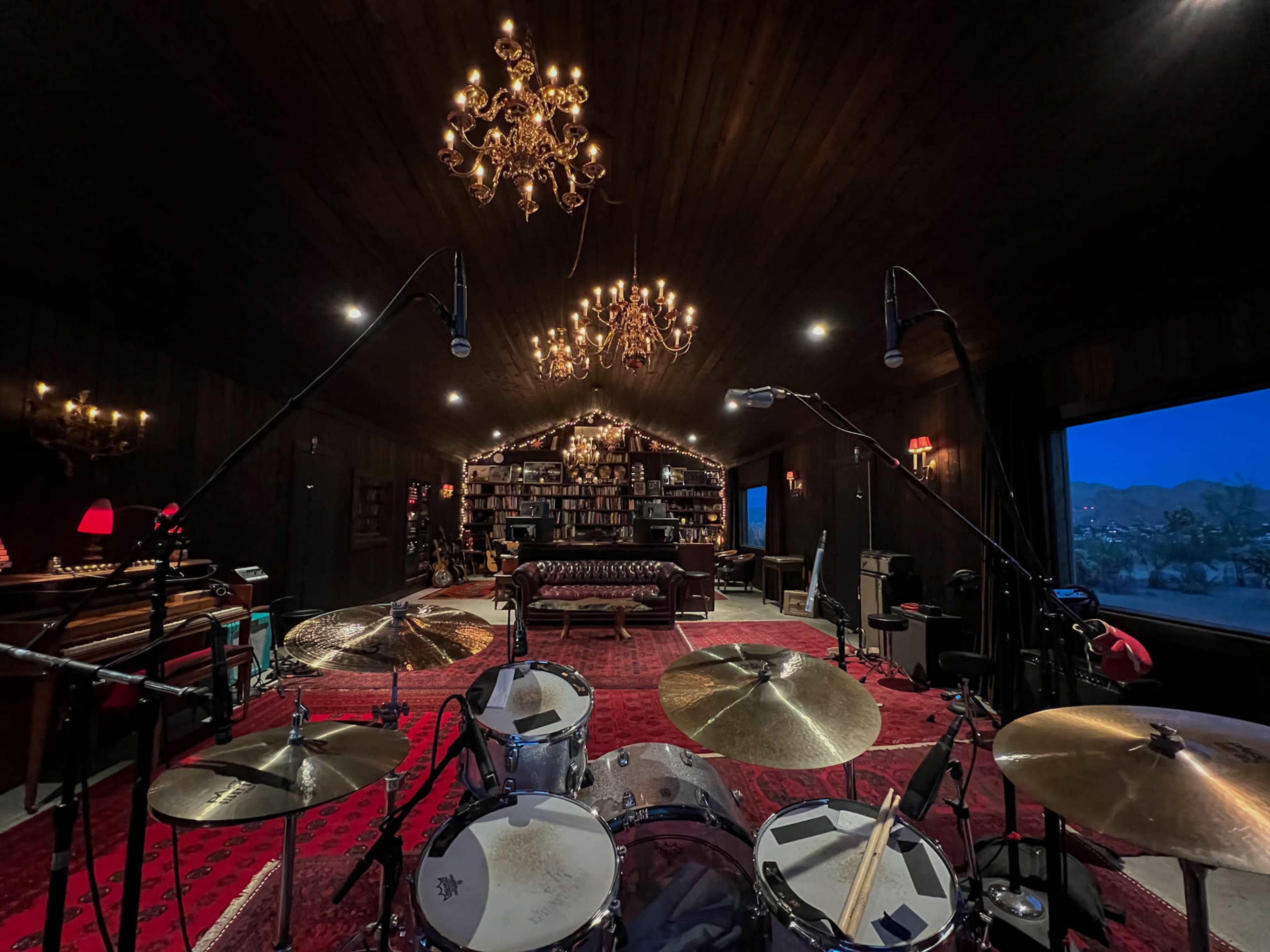 The image shows a dimly lit music studio featuring a drum set in the foreground, an ornate chandelier overhead, and a cozy seating area backed by a wall of bookshelves.