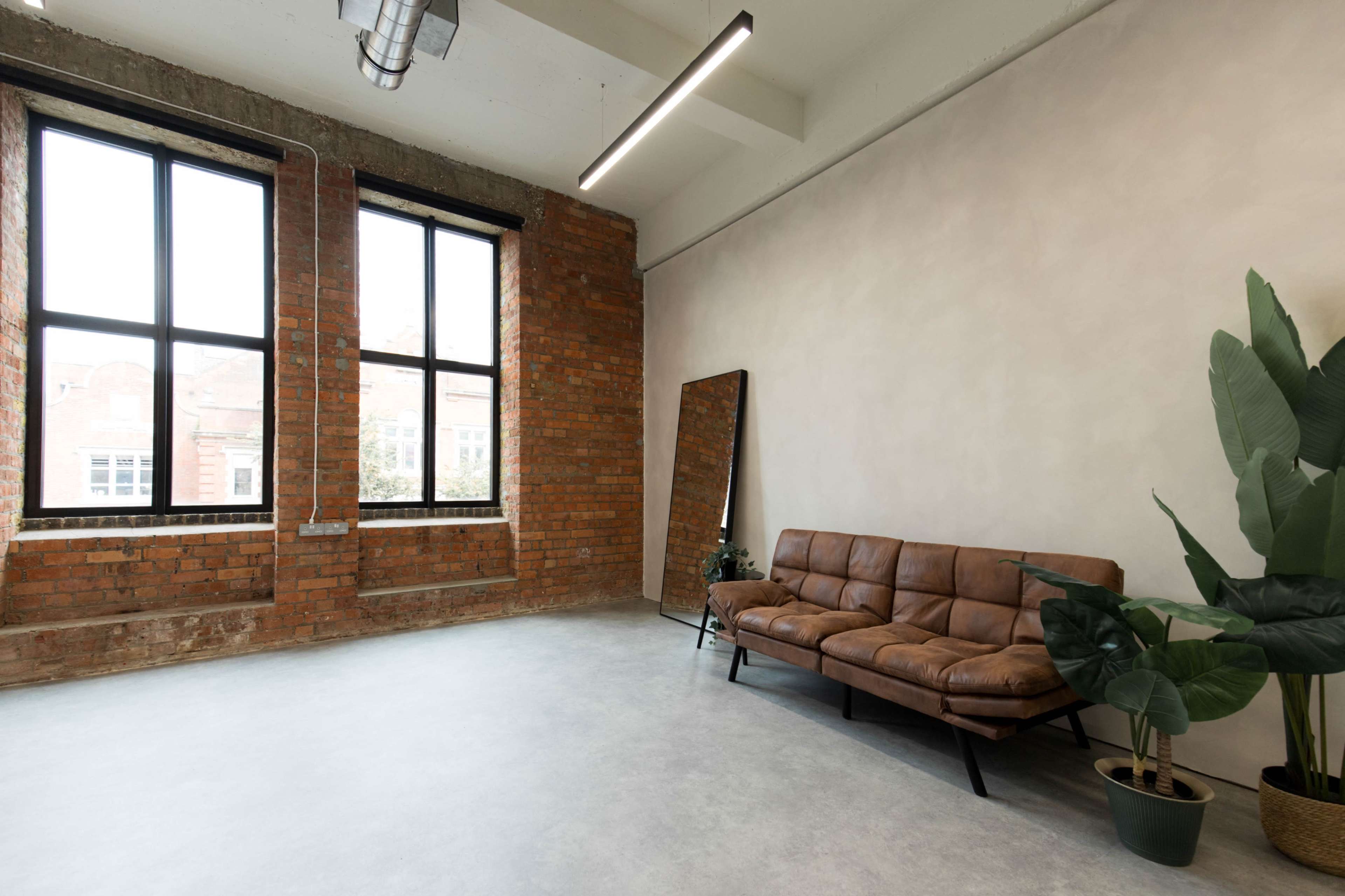 The image shows a modern, minimalist interior space with exposed brick walls, large windows, a brown leather sofa, a floor mirror, and a potted plant.