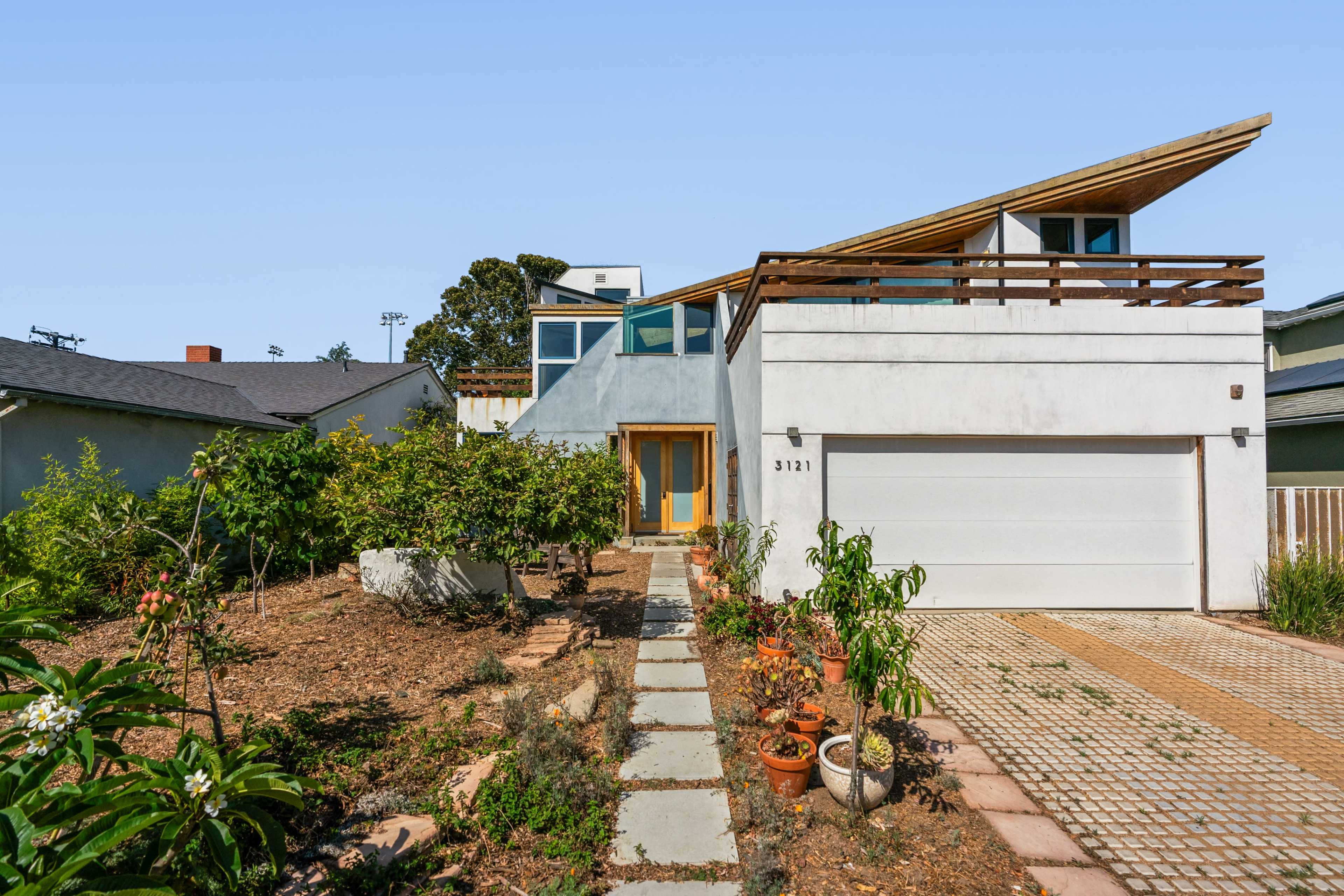 The image shows a modern two-story house with a sloped roof, a landscaped front yard, and a driveway leading to a garage.