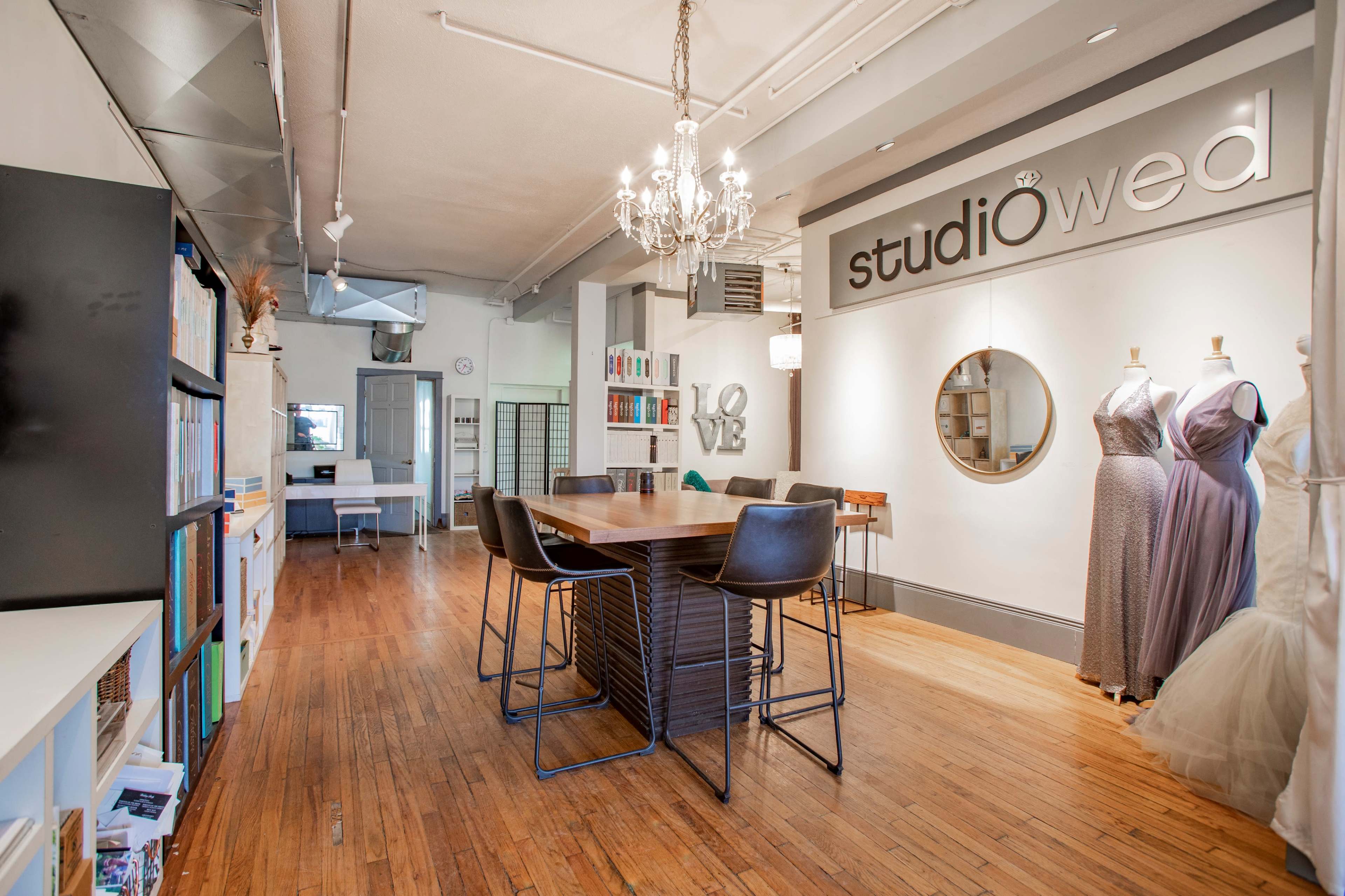 A modern bridal studio with a central wooden dining table surrounded by black chairs, displaying elegant wedding dresses on mannequins and bookshelves lining the walls.
