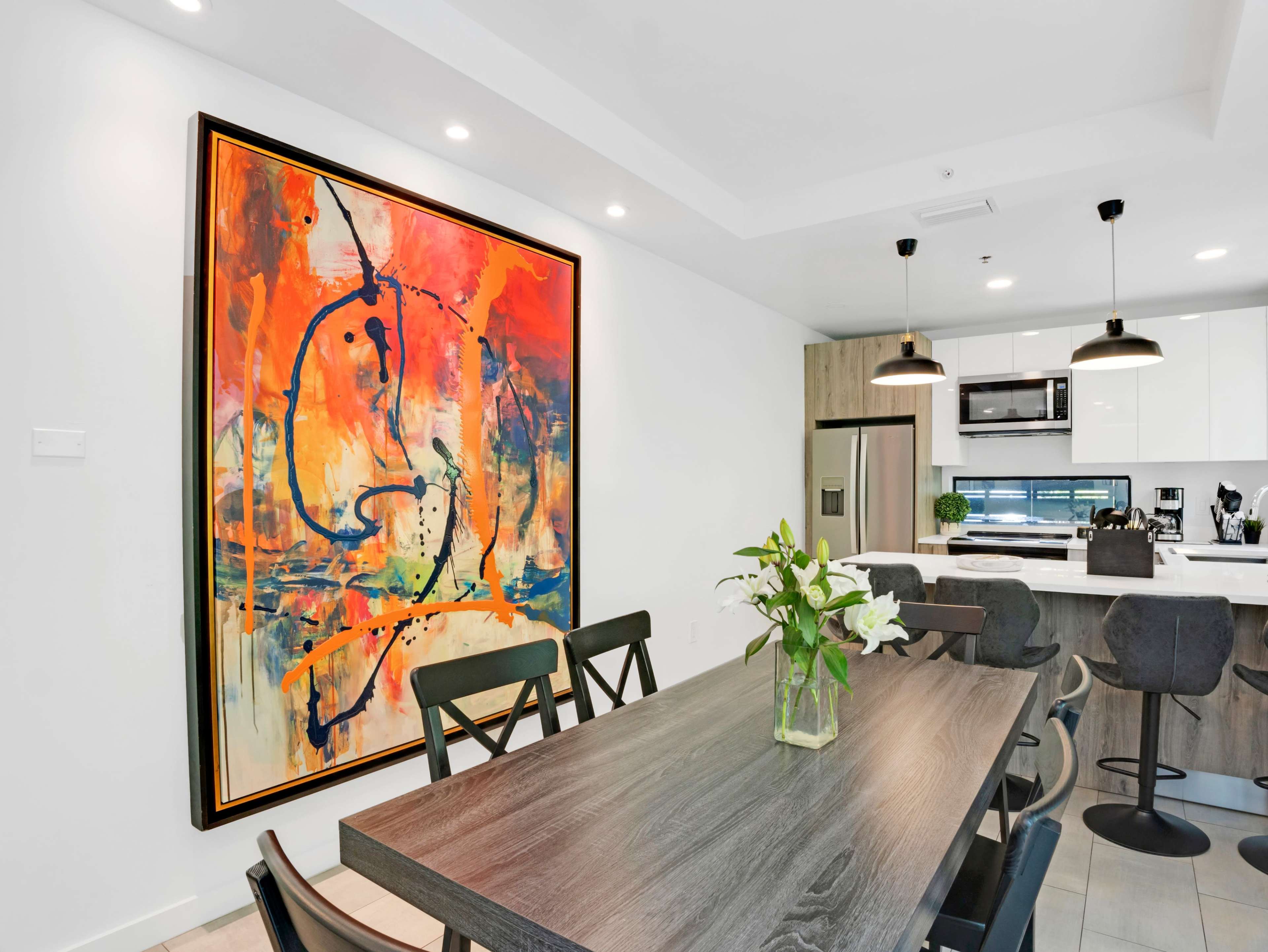 A dining area features a large abstract painting on the wall above a wooden table surrounded by black chairs, with a kitchen visible in the background.