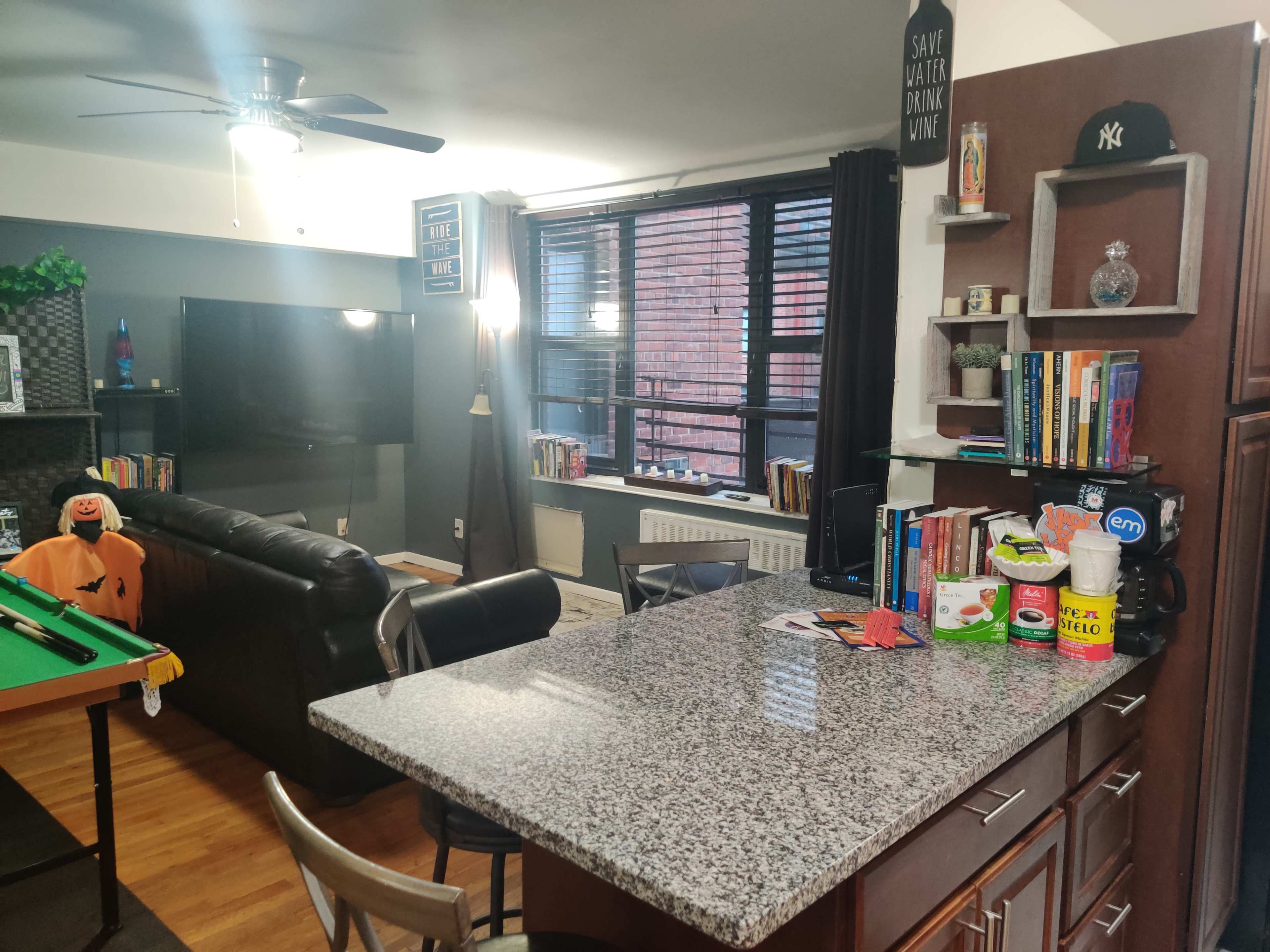 An open living space with a kitchen counter, a black sofa, a television, and a bookshelf against a wall with large windows.