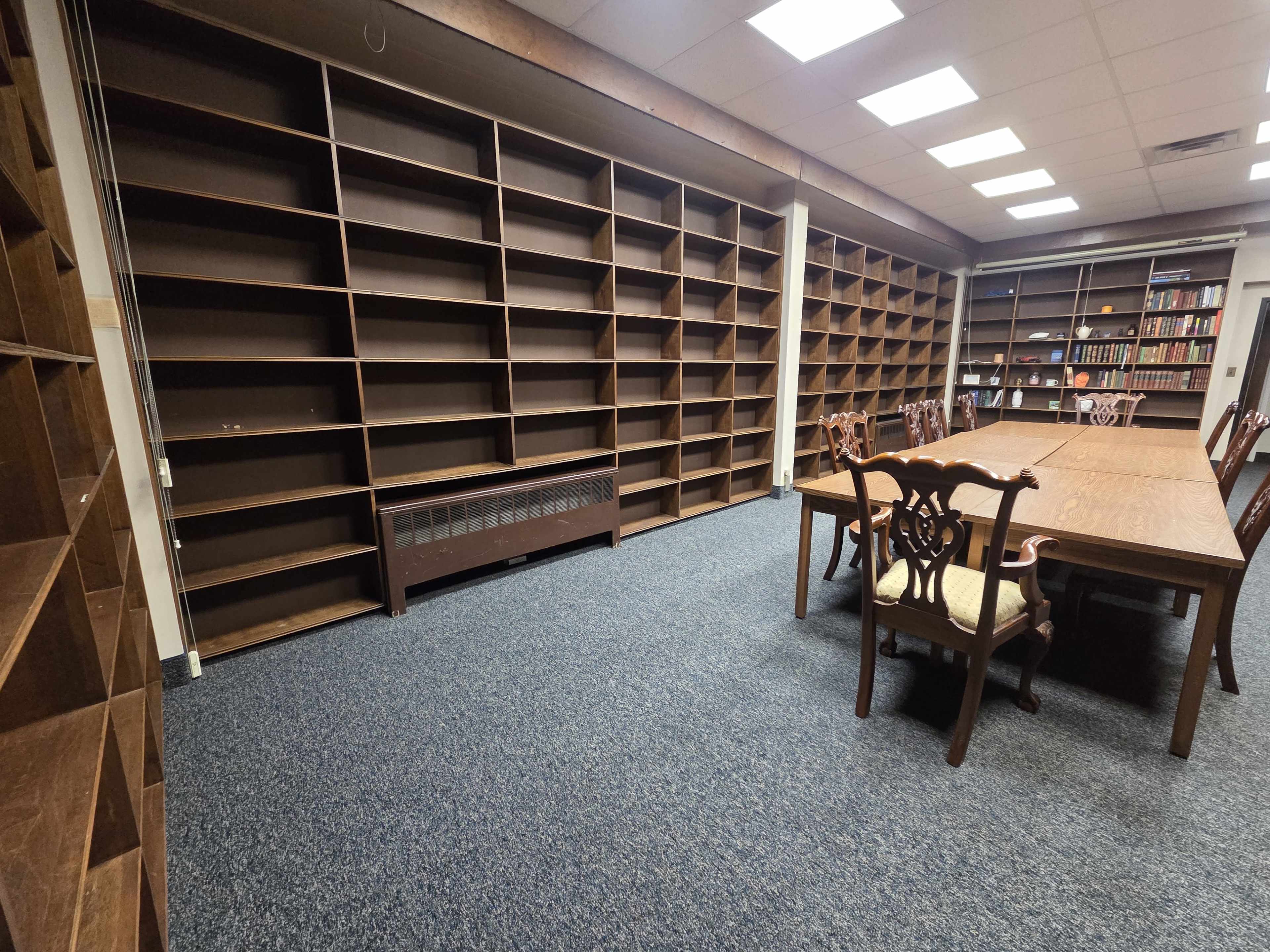 The image shows a room with a large wooden table surrounded by chairs, and two walls lined with empty bookshelves.