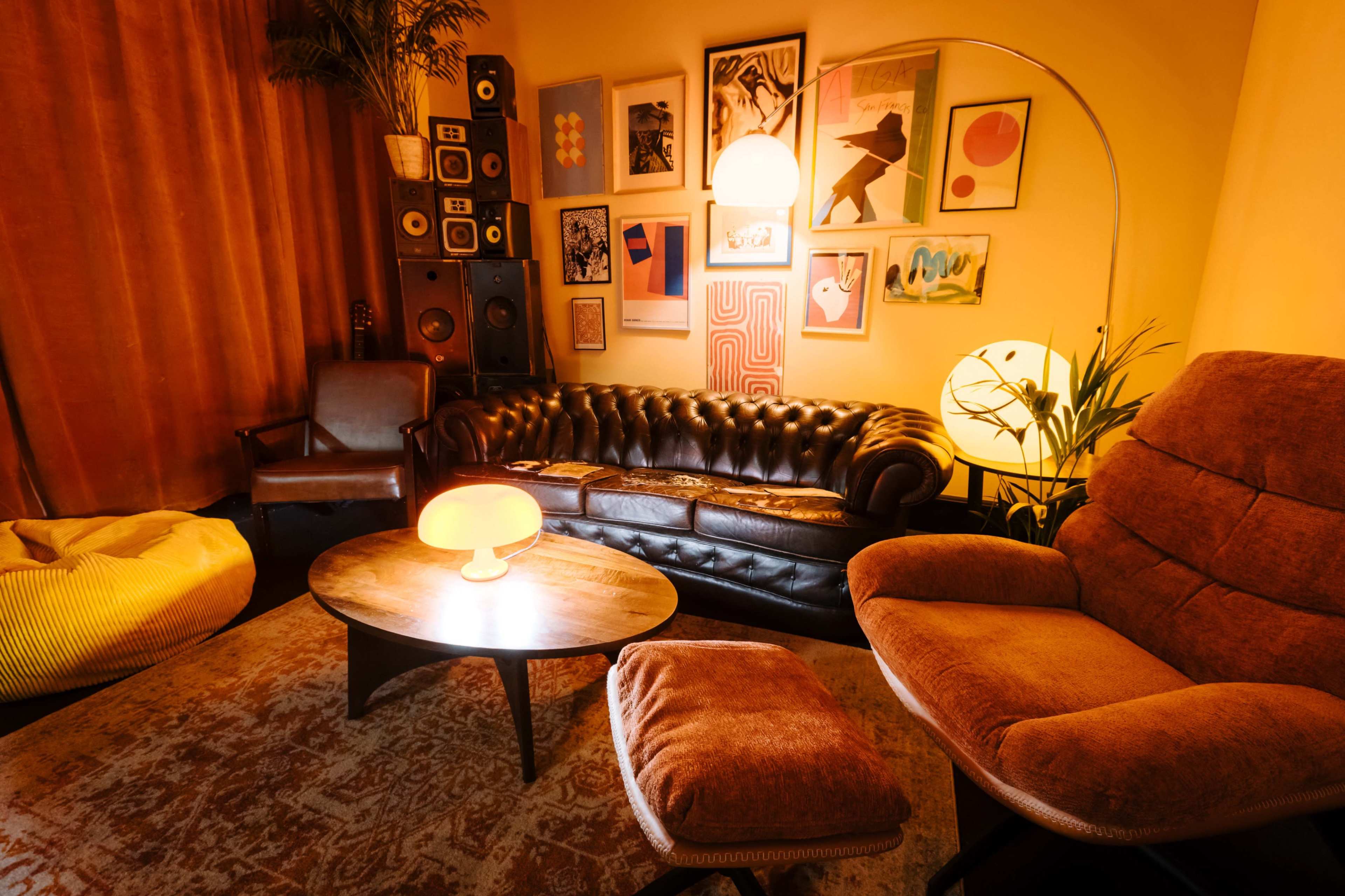 A cozy living room features a leather chesterfield sofa, a round wooden coffee table, and various wall art, illuminated by warm lighting.