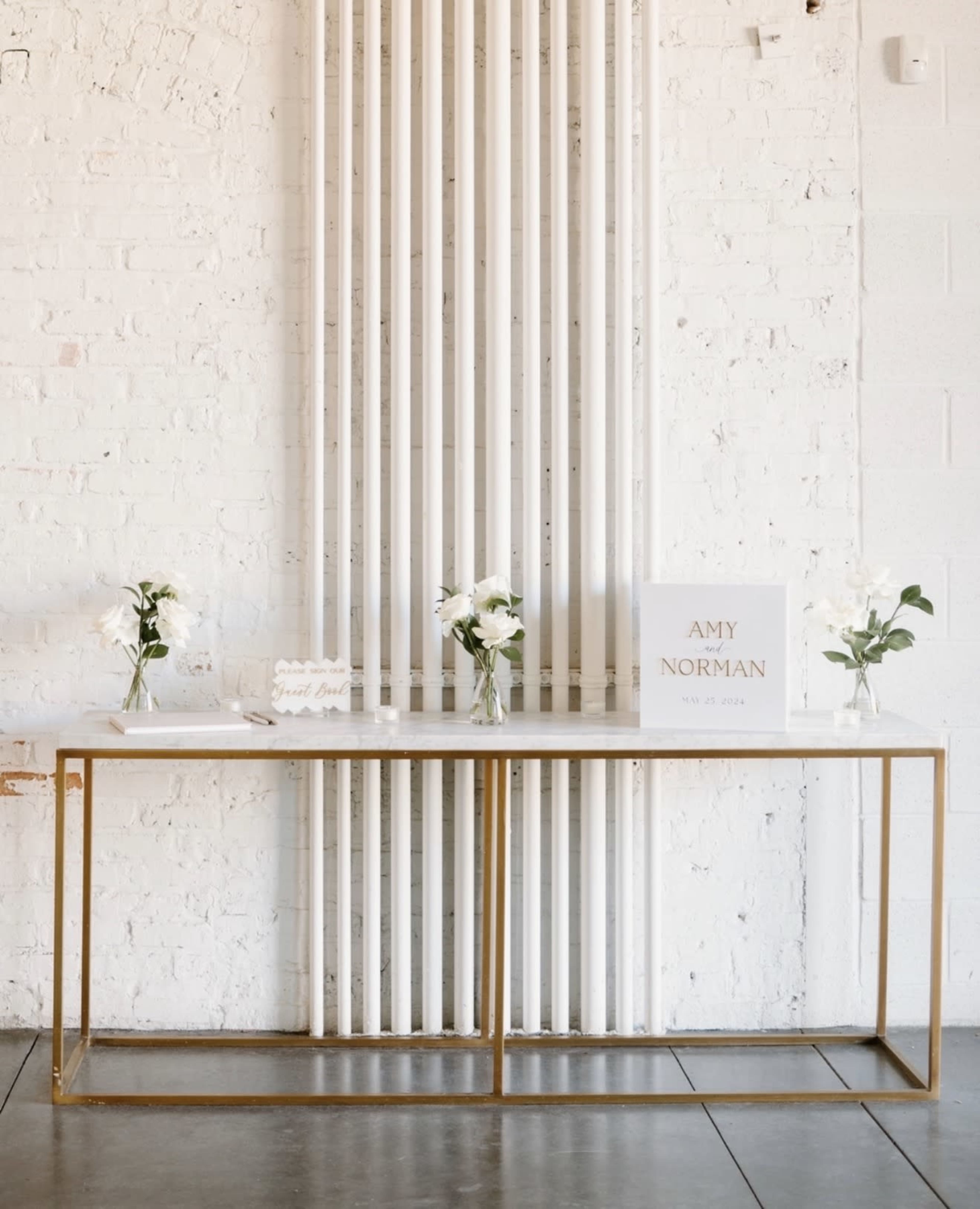 A long, metal-framed table with a white top displays two vases of flowers and a sign against a backdrop of white brick and vertical white piping.