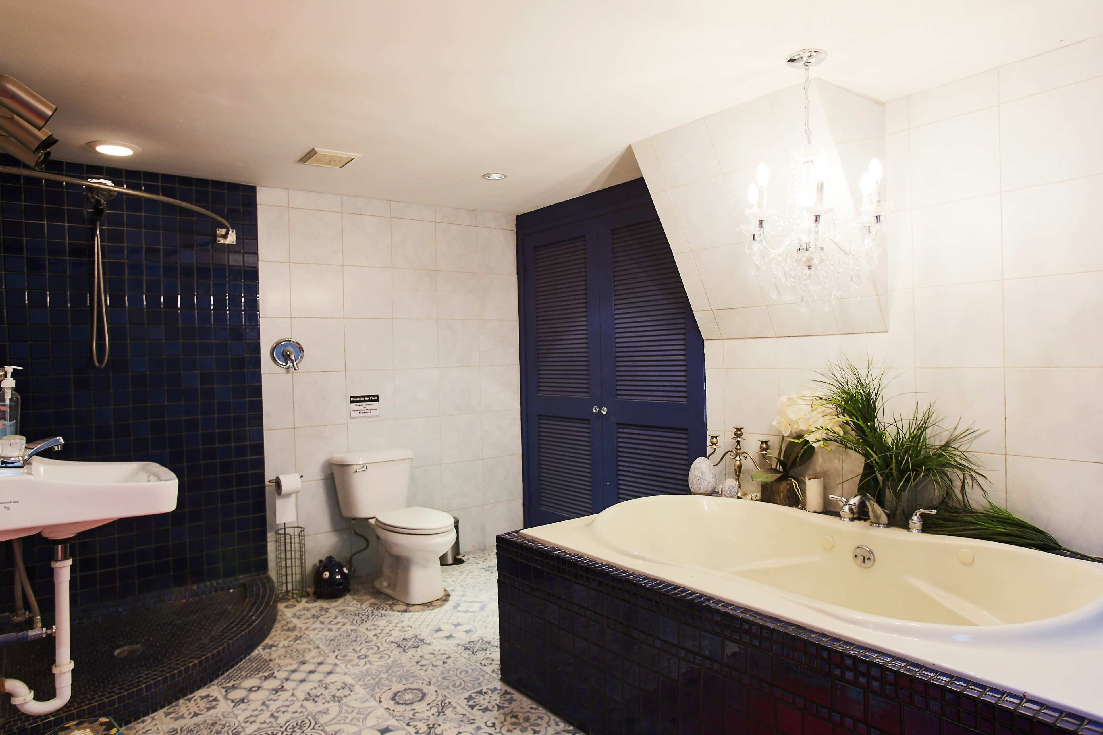 A bathroom featuring a shower area, a white toilet, a double sink with a blue tiled wall, and a large bathtub, all set against light-colored tiles and illuminated by a chandelier.