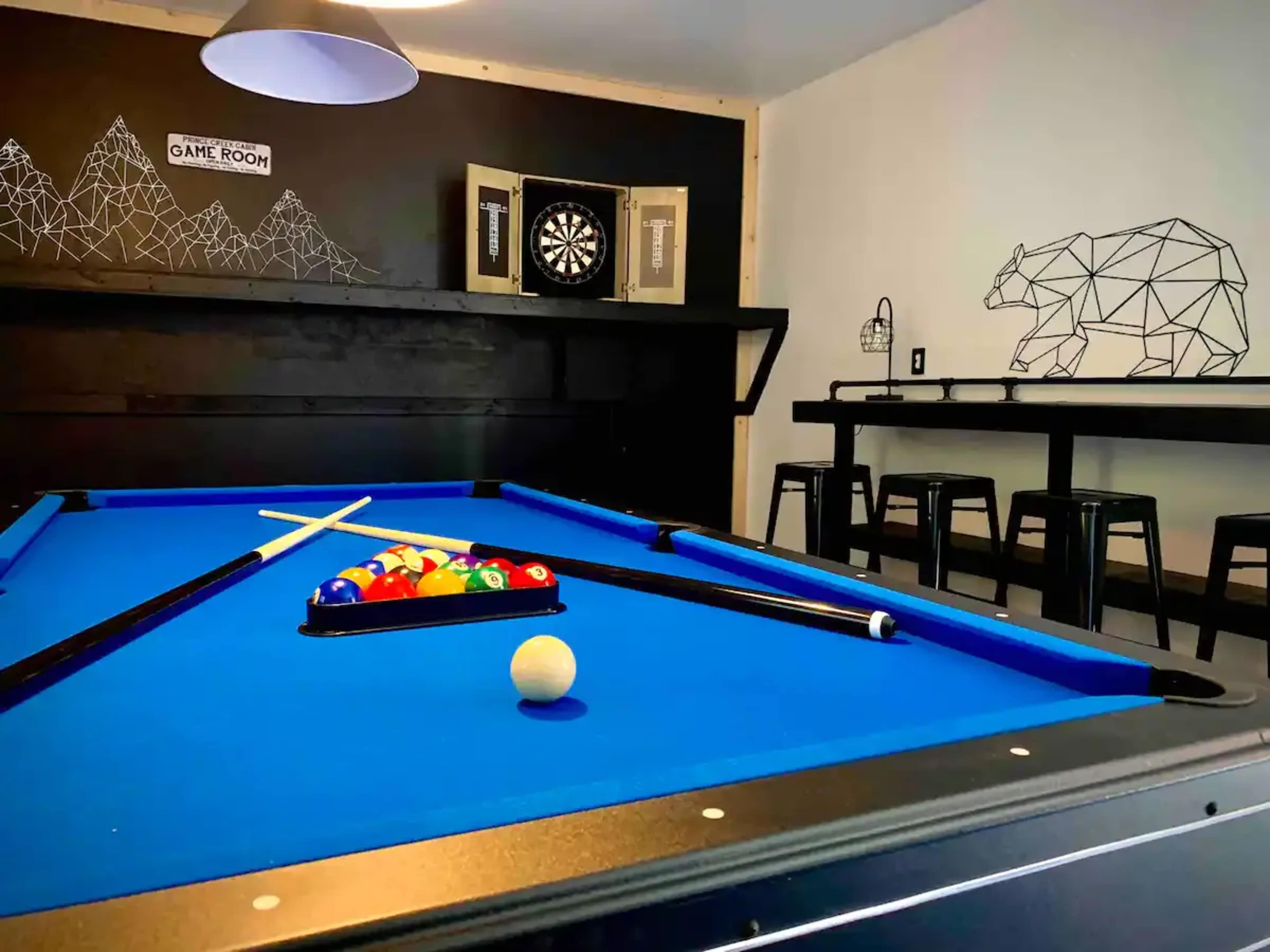 The image shows a game room featuring a blue pool table with balls arranged in a rack, a dartboard on the wall, and a bar-height table with stools.