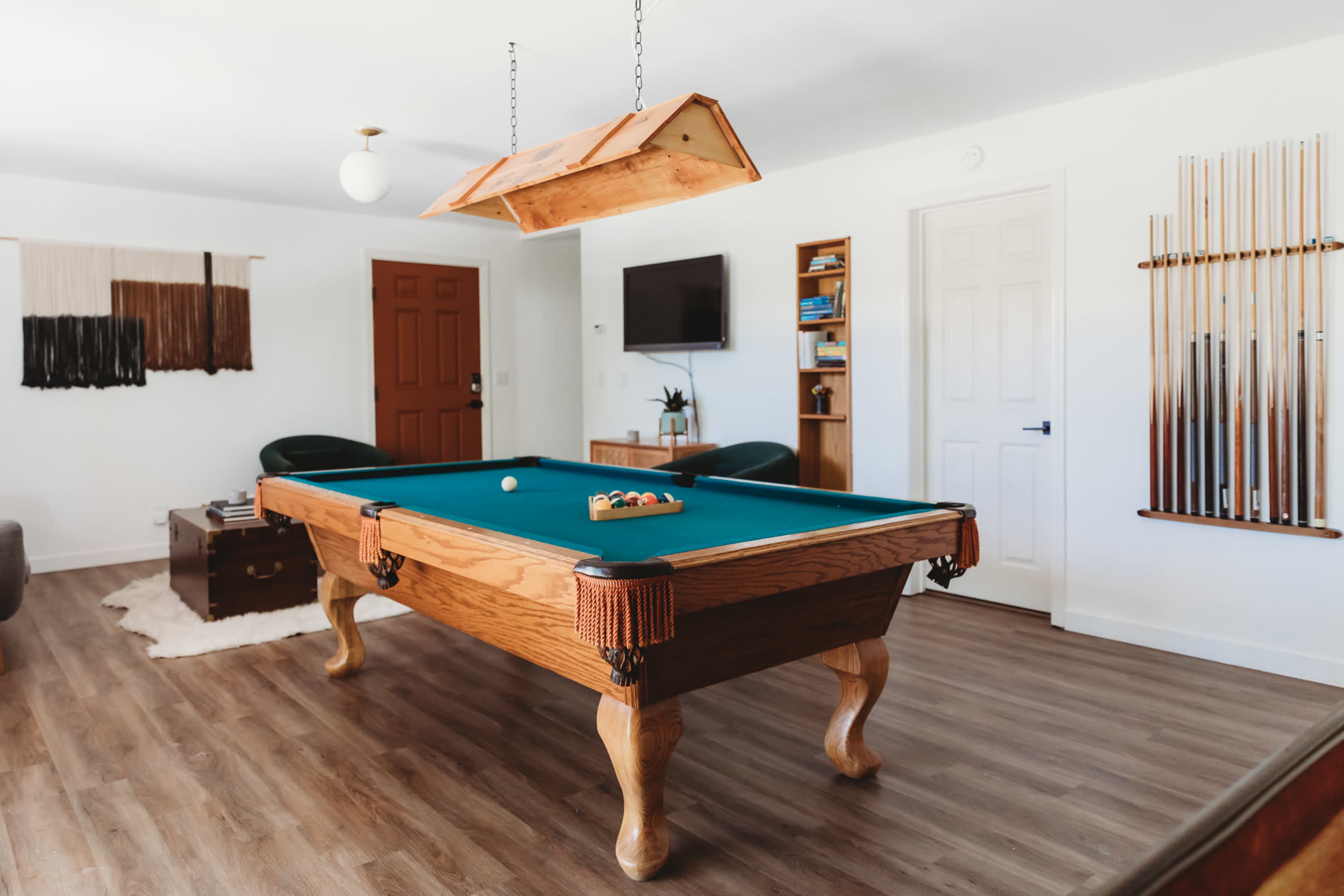 A brightly lit game room featuring a wooden pool table with a green felt surface, surrounded by leather chairs and various pool cues displayed on the wall.