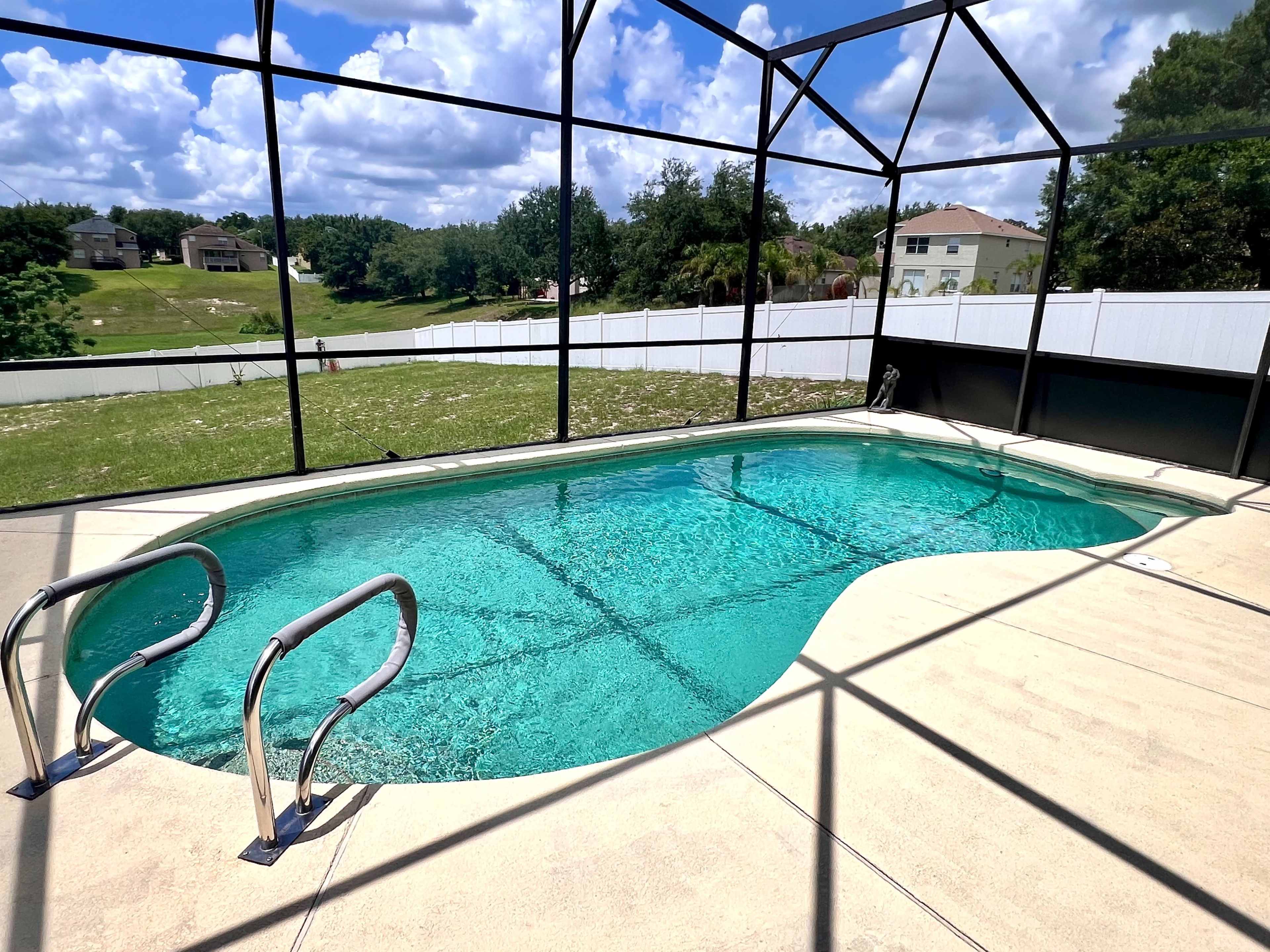 The image shows a rectangular swimming pool with a light blue water surface, surrounded by a screened-in enclosure and grassy landscaping in the background.