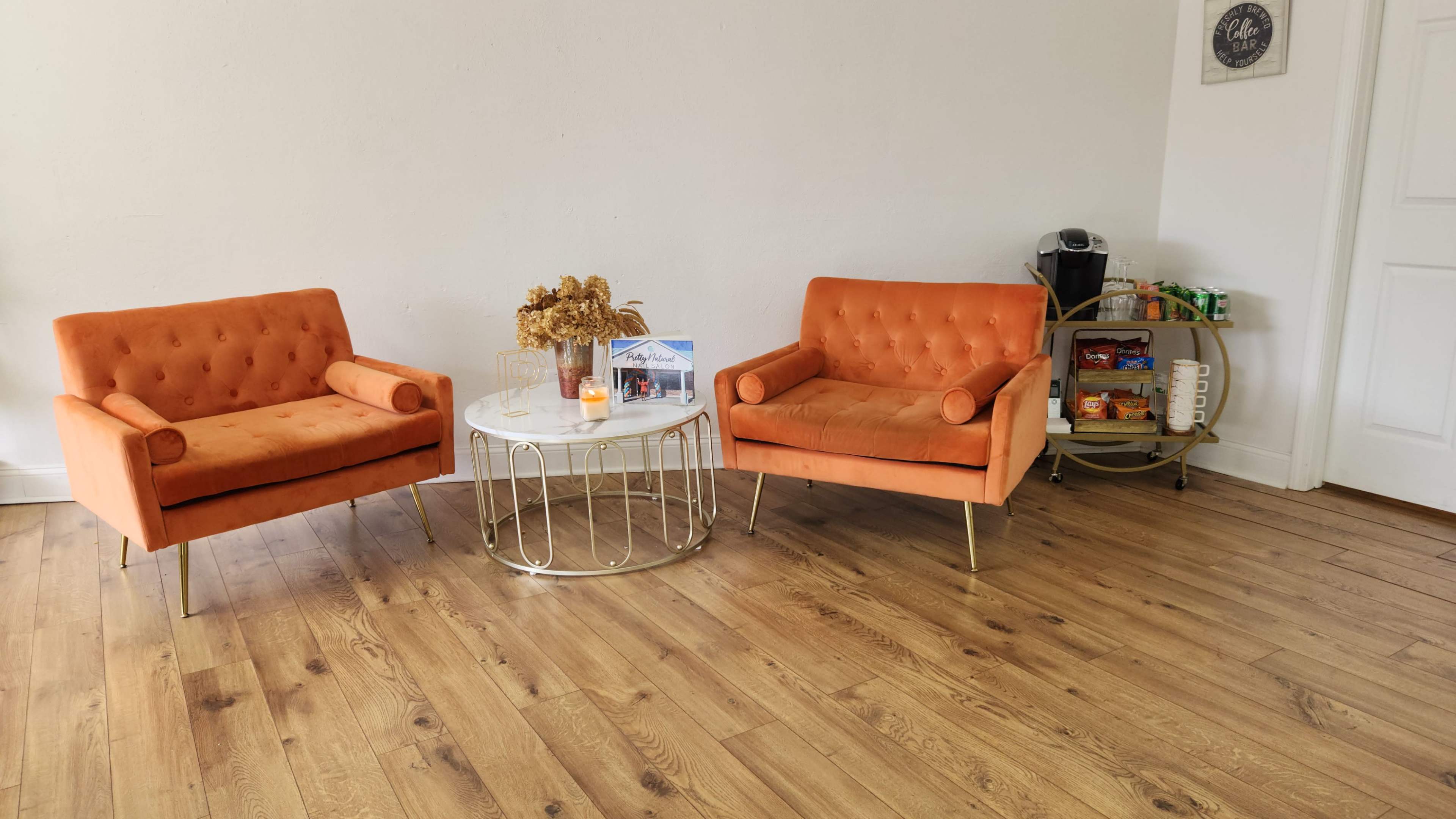 The image features two orange velvet chairs facing a round coffee table, which is adorned with dried flowers, in a room with light wooden flooring and a small cart filled with snacks on the side.