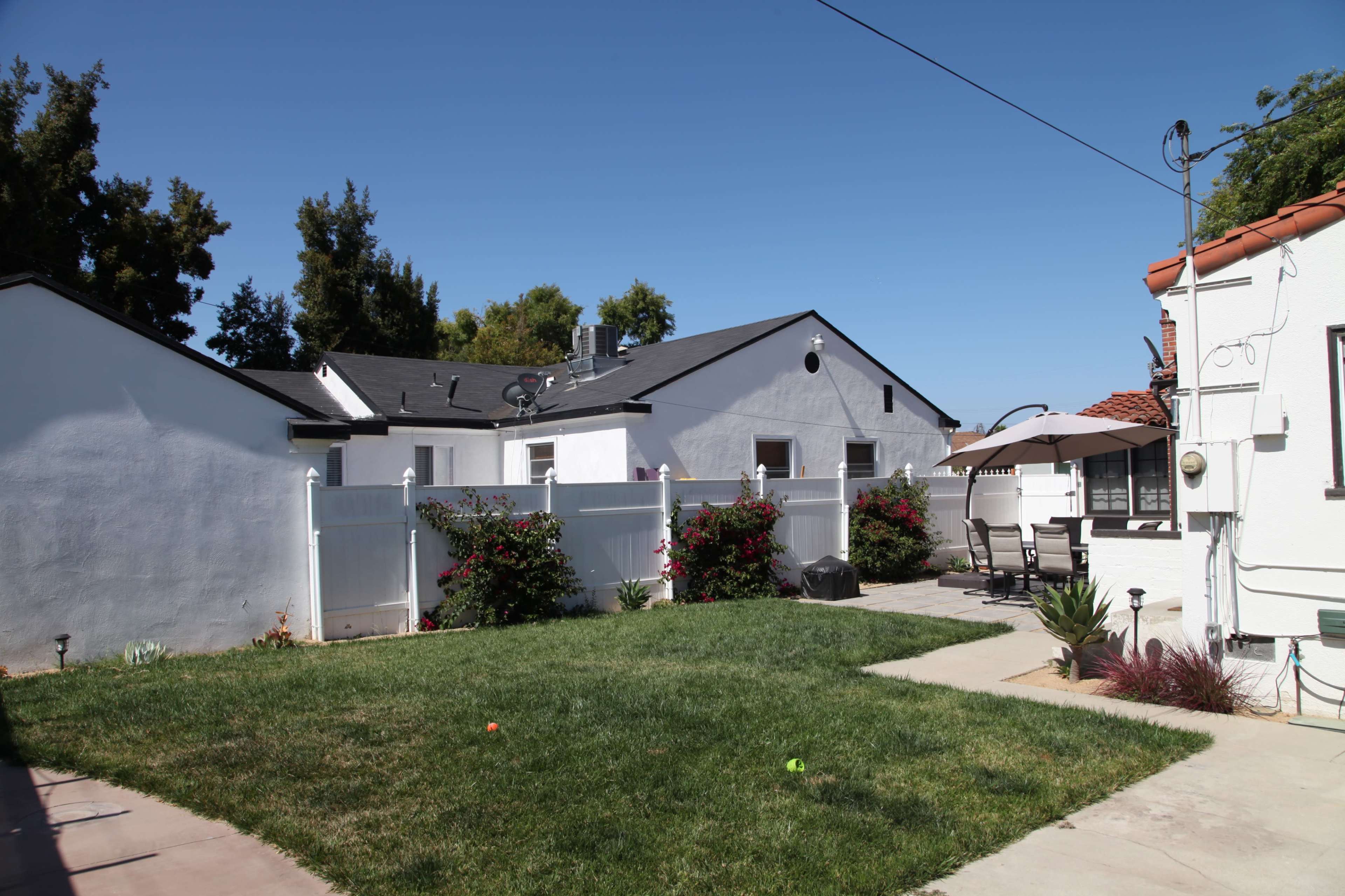 A backyard with a green lawn, bordered by white fences and blooming bushes, alongside a patio area with outdoor furniture and an umbrella.