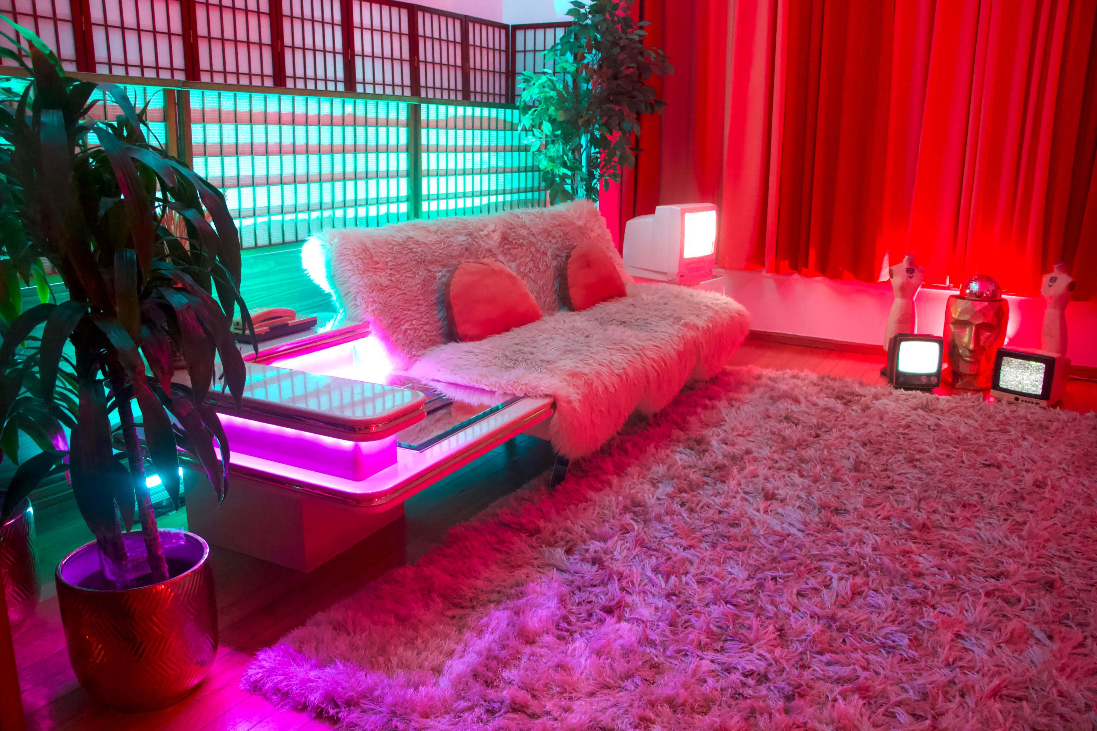 A brightly colored retro-styled living room featuring a plush sofa, a large rug, and various decorative elements illuminated with vibrant lighting.