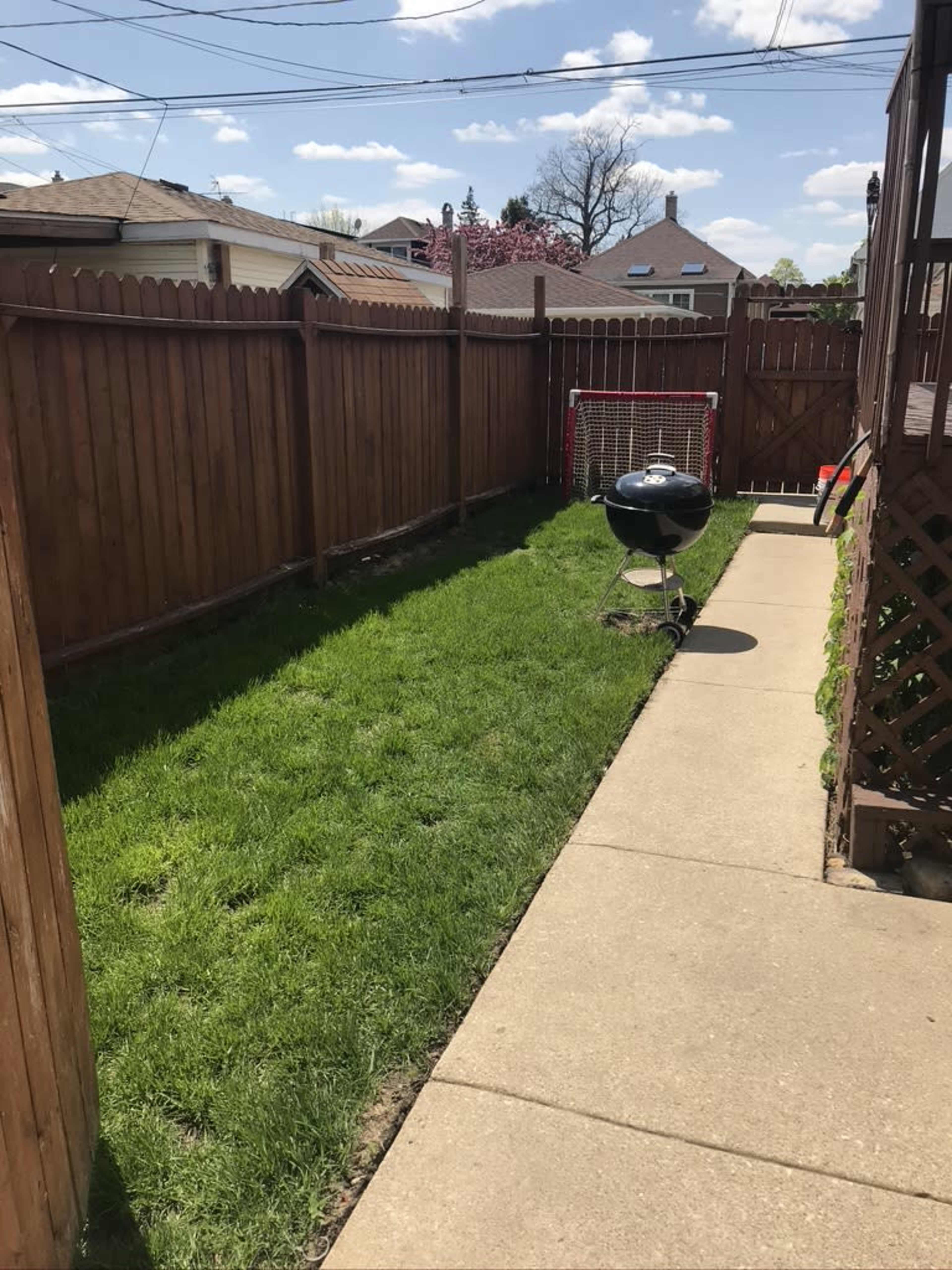 The image shows a narrow backyard with a green lawn bordered by wooden fencing and a charcoal grill on a concrete path.