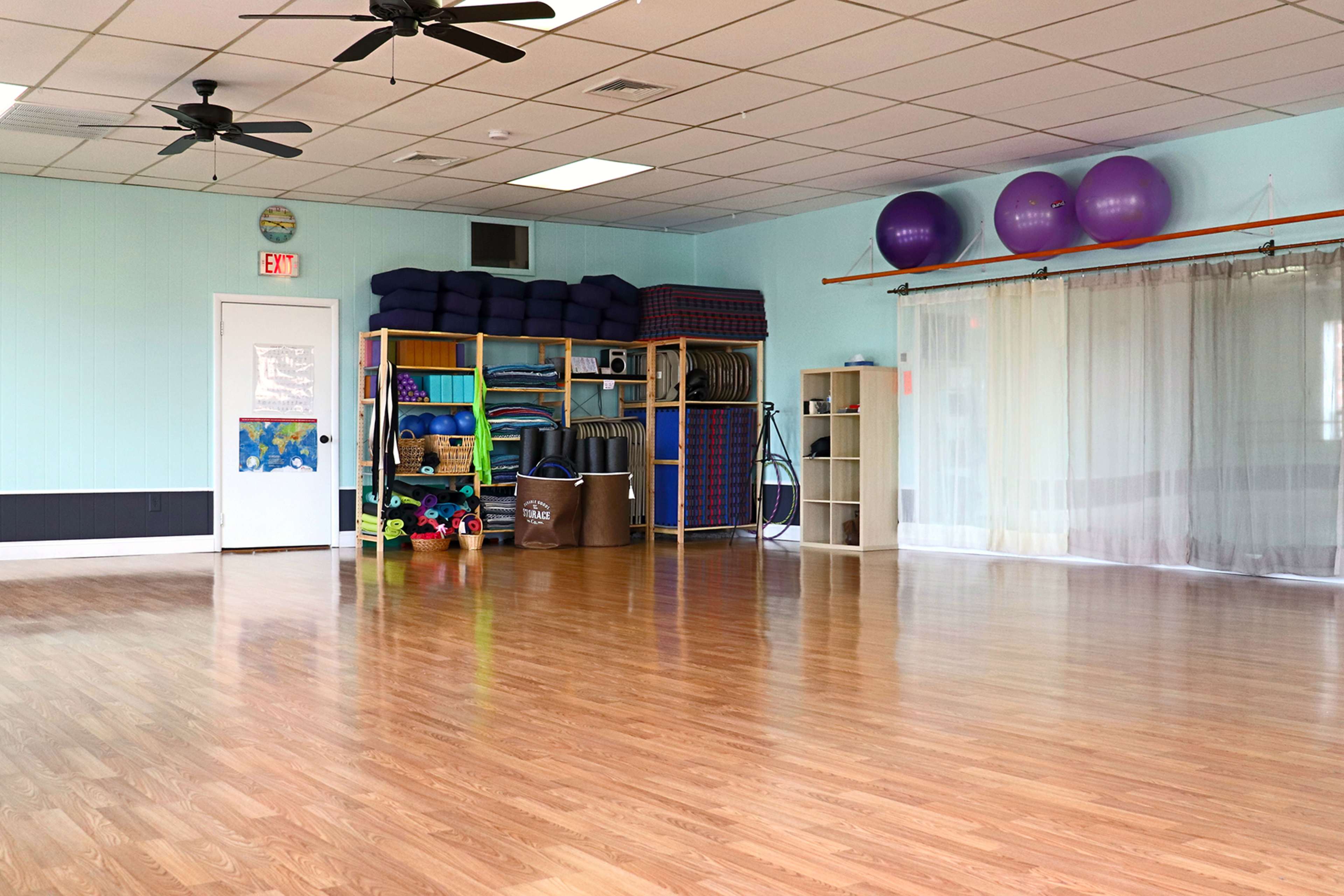 An empty fitness or multipurpose room with wooden flooring, shelves holding exercise equipment, and a curtain-covered area at one end.