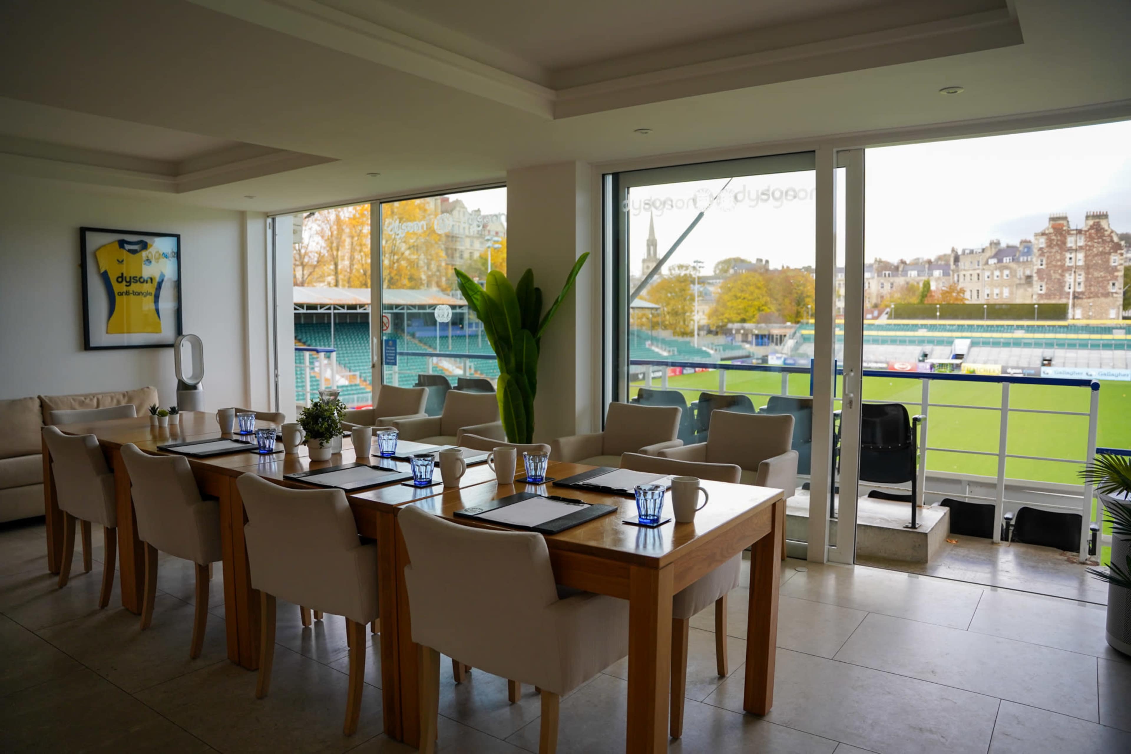 A bright dining area with a long wooden table set for a meal, overlooking a sports field through large glass windows.