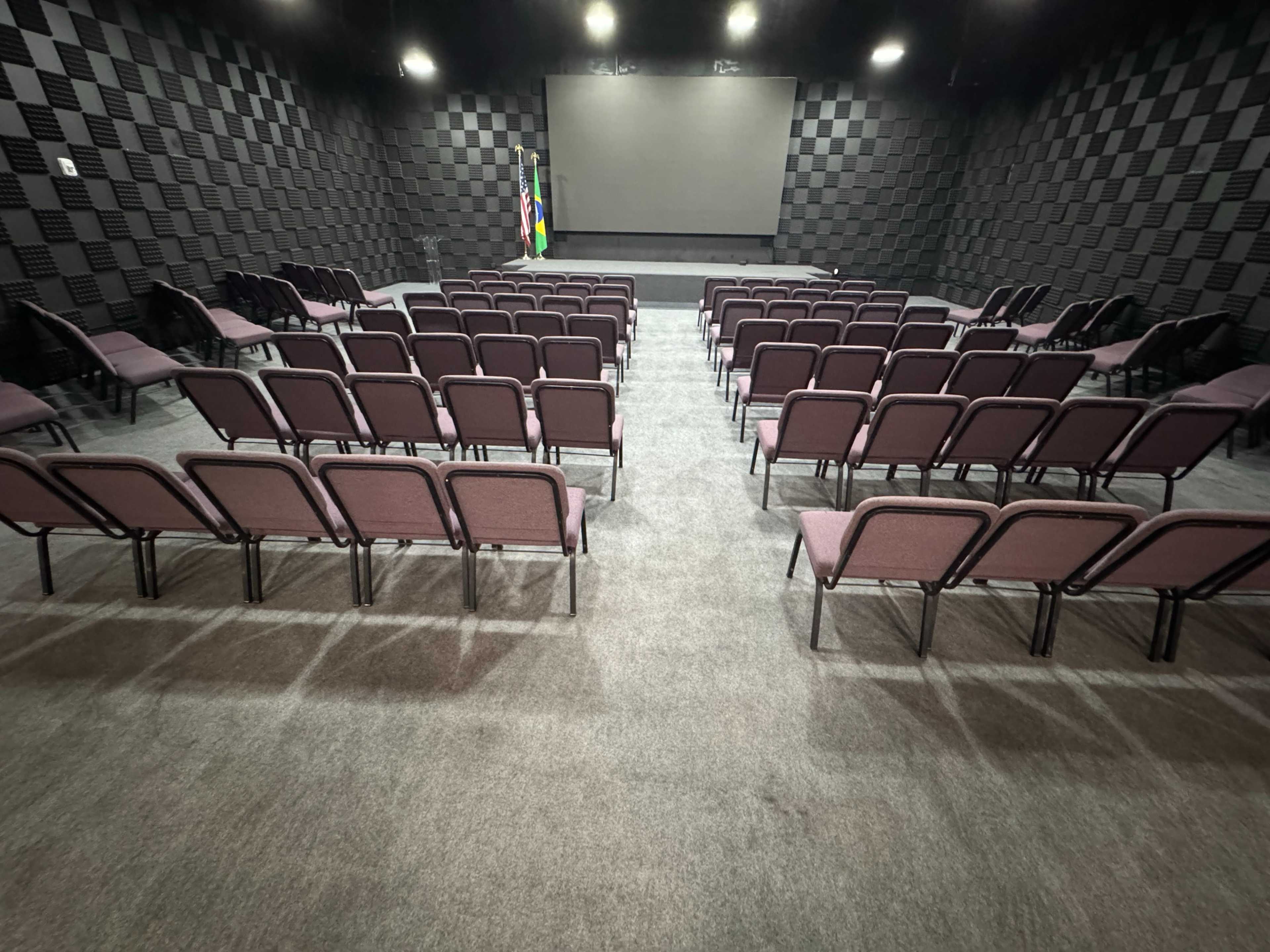 The image shows an empty auditorium with rows of maroon chairs arranged in front of a large screen and dark checkered walls.