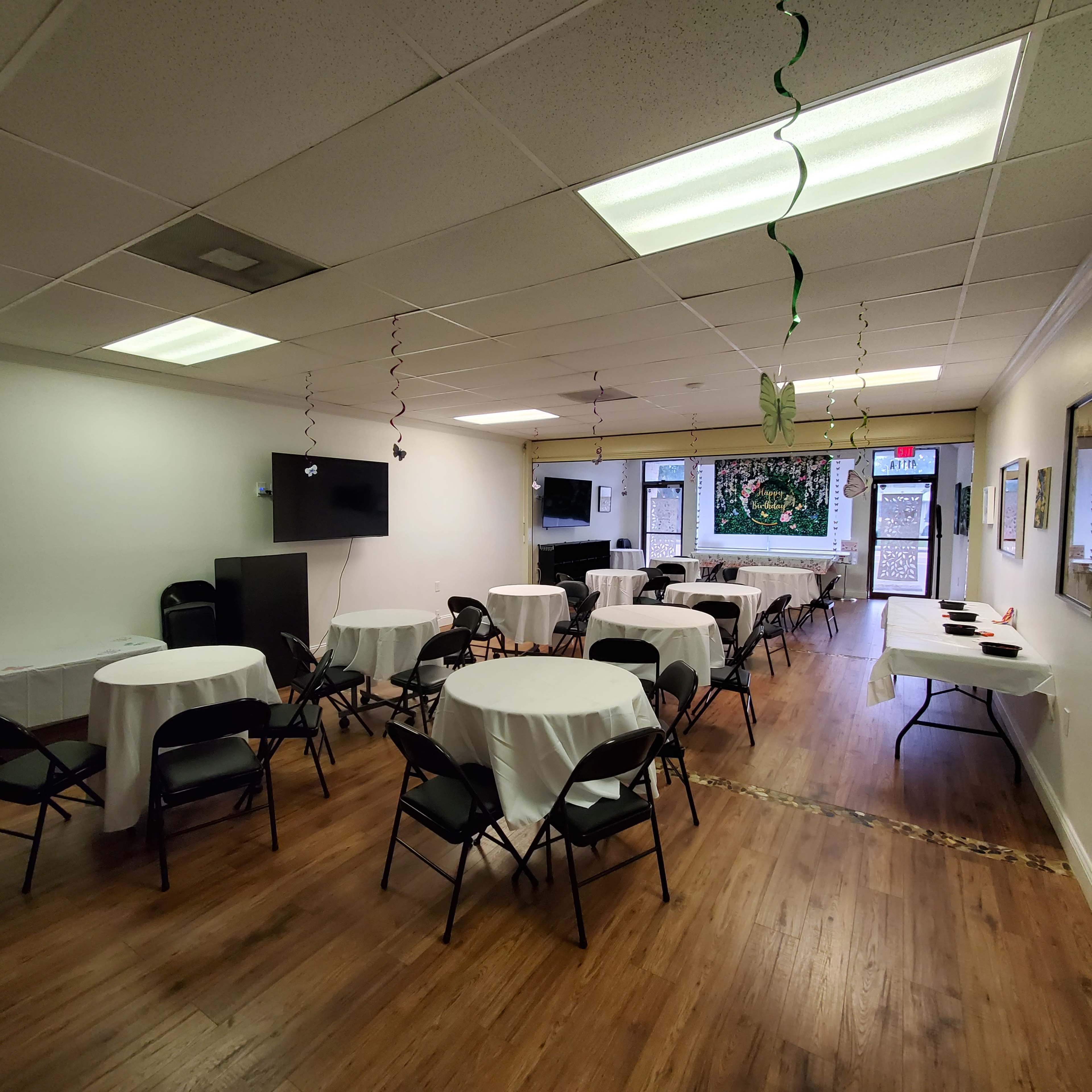 The image shows a large room set up with several round tables covered in white tablecloths and black chairs arranged around them, along with decorations hanging from the ceiling.