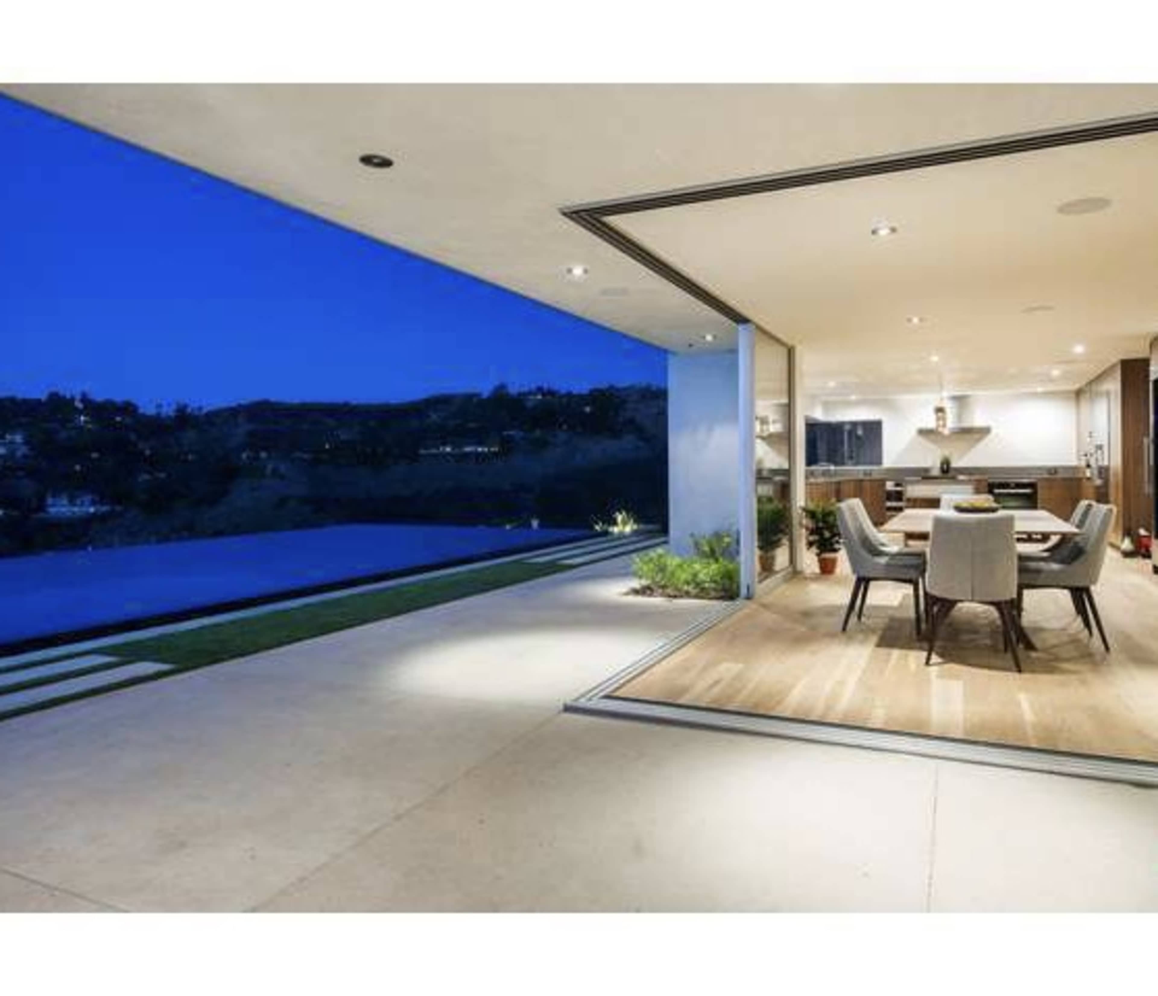 A modern dining area opens up to an outdoor space with a pool and hillside view at dusk.
