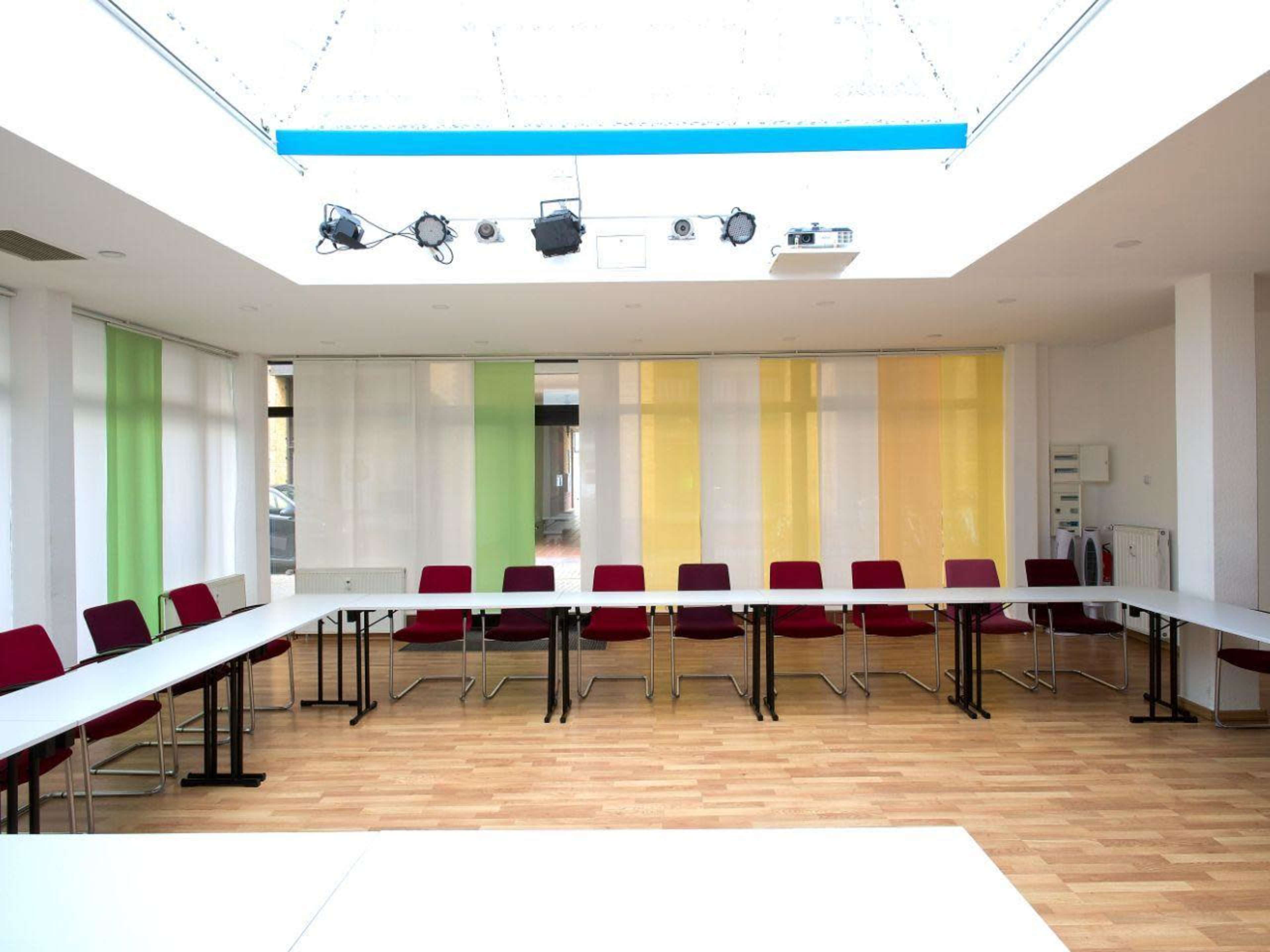A modern conference room features a long table surrounded by red chairs, with colorful translucent curtains letting in natural light.