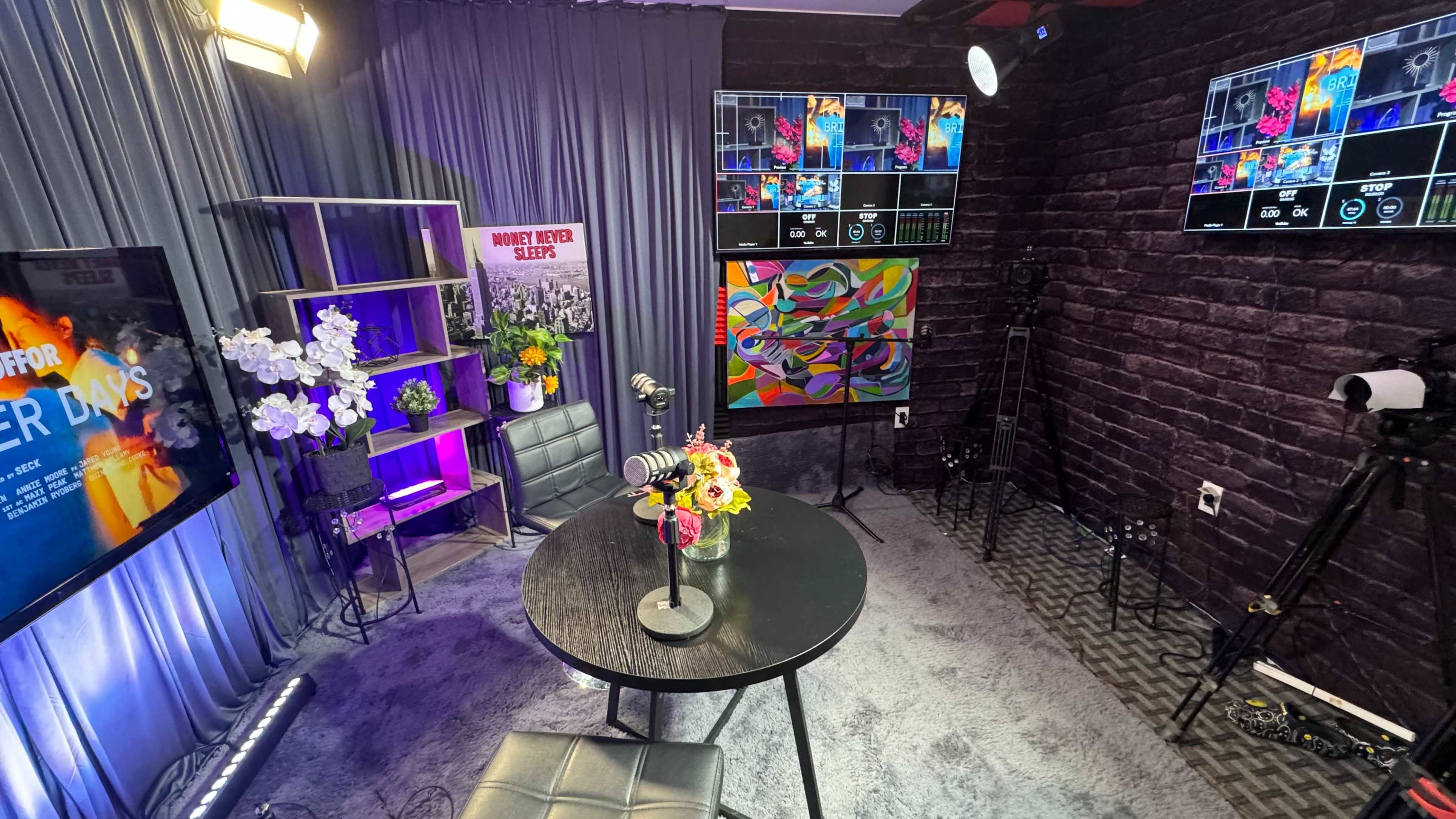 The image shows a modern studio setup with a round table, a microphone in the center, vibrant artwork on the wall, and multiple screens displaying graphics.
