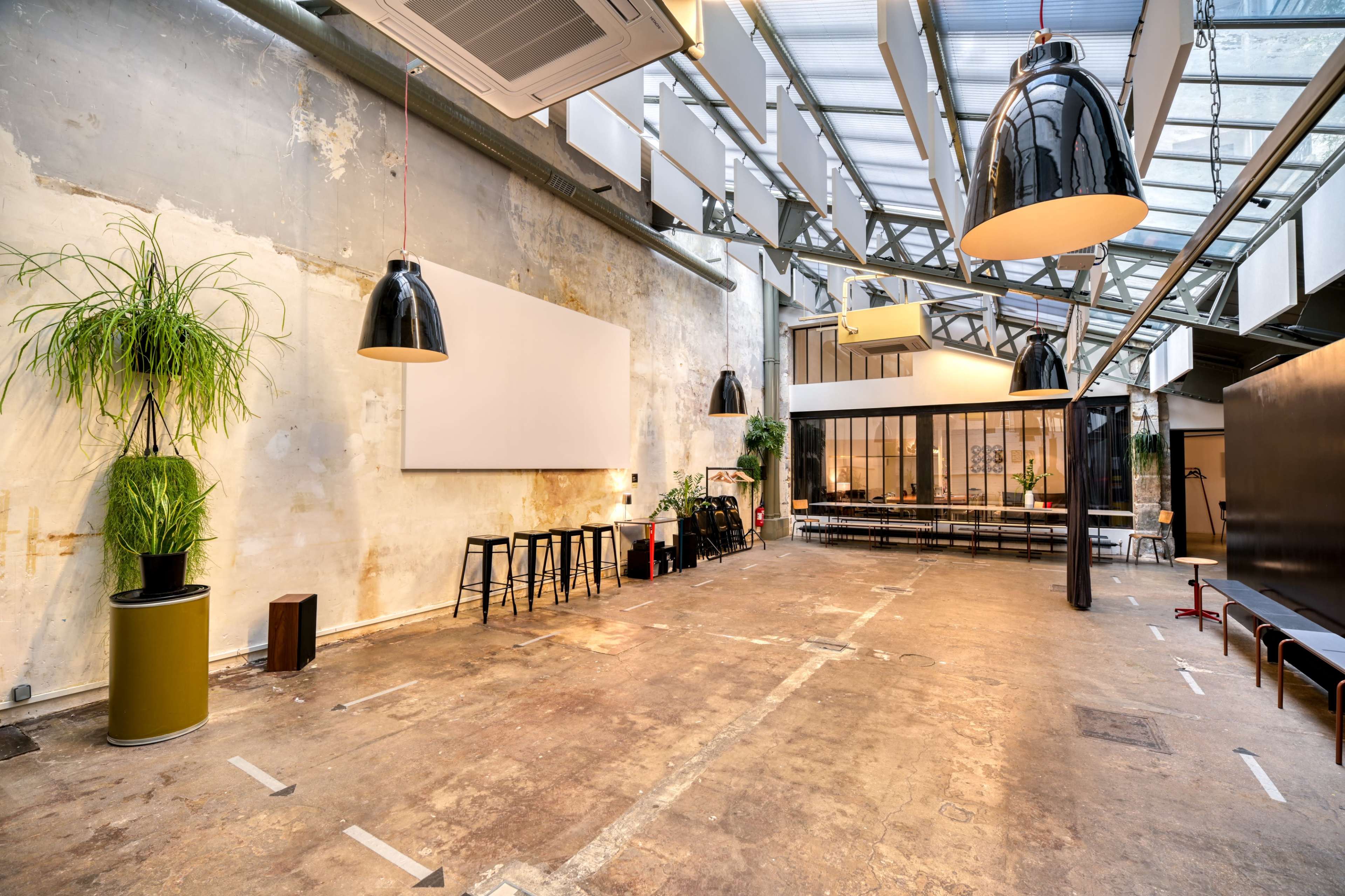The image shows an industrial-style, spacious room with a bright glass ceiling, wooden floors, and minimalist furnishings including a large screen and black stools.