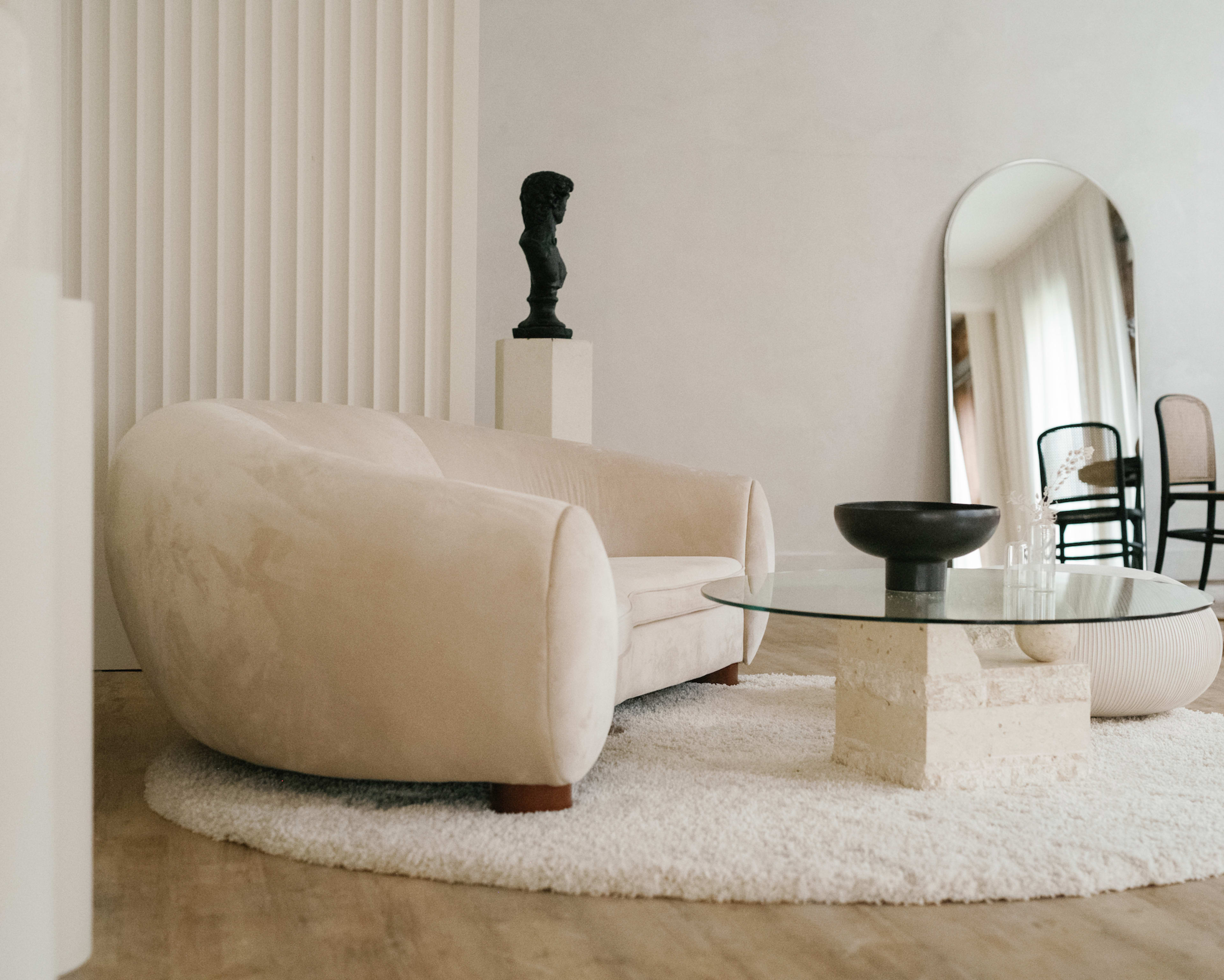 A plush beige sofa sits on a textured rug beside a round glass coffee table, with a sculptural bust and a mirror in the background.