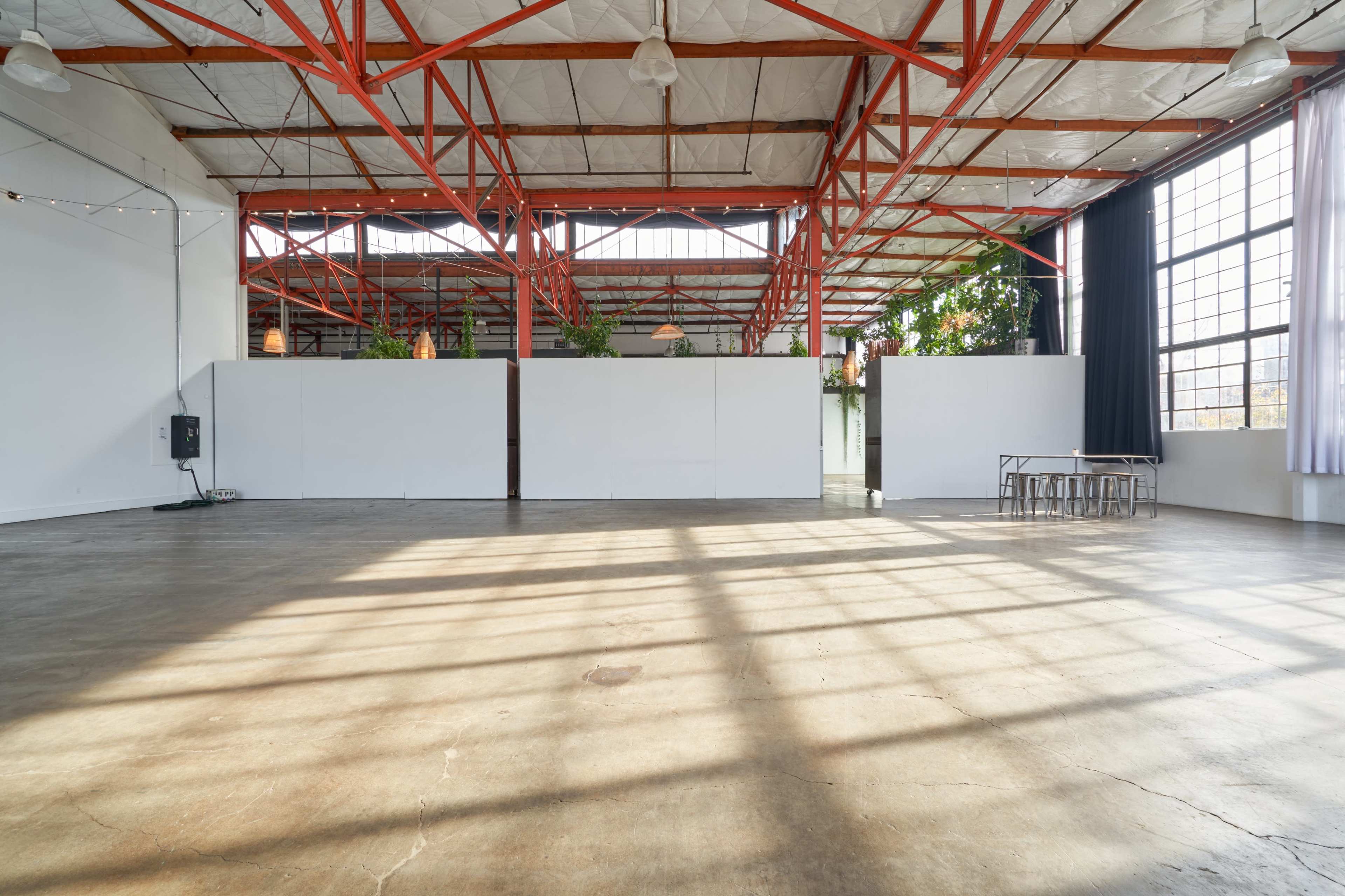 An empty industrial space with high ceilings, large windows, and exposed red steel beams, featuring concrete flooring and white partition walls.