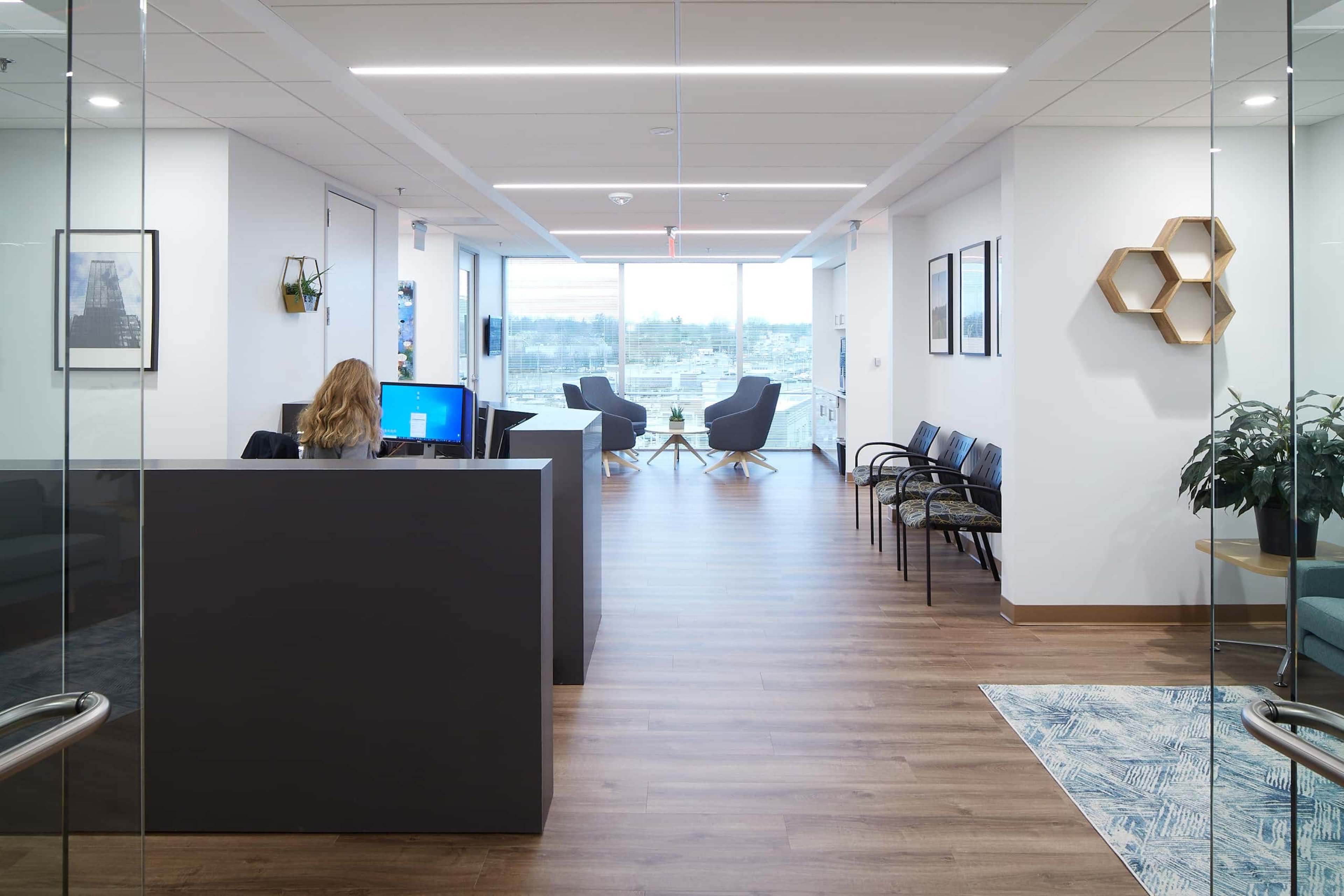 A modern office reception area features a desk with a computer, a row of waiting chairs, and large windows overlooking a cityscape.