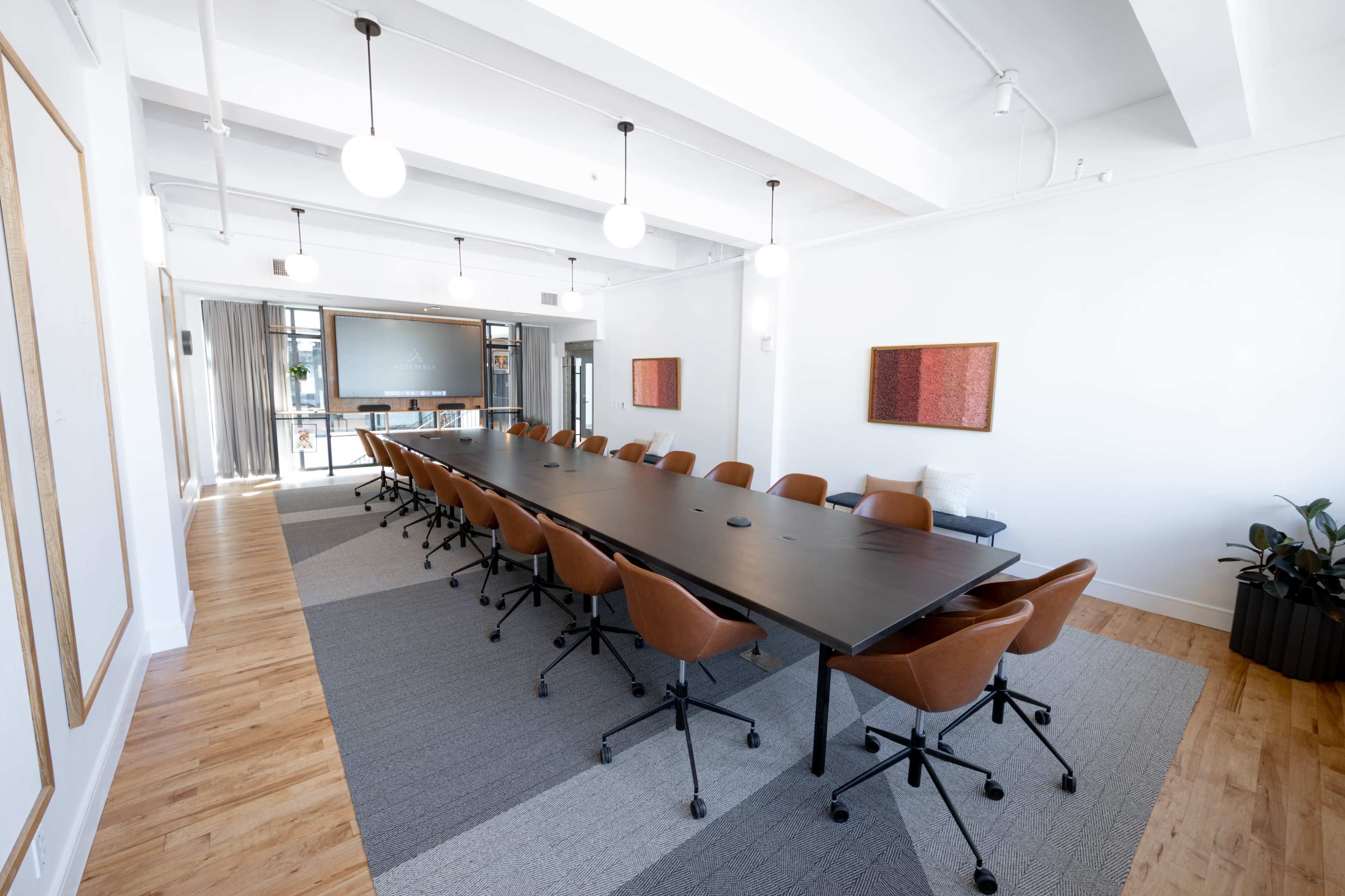 A long conference table with brown chairs is set up in a well-lit meeting room featuring large windows and modern decor.