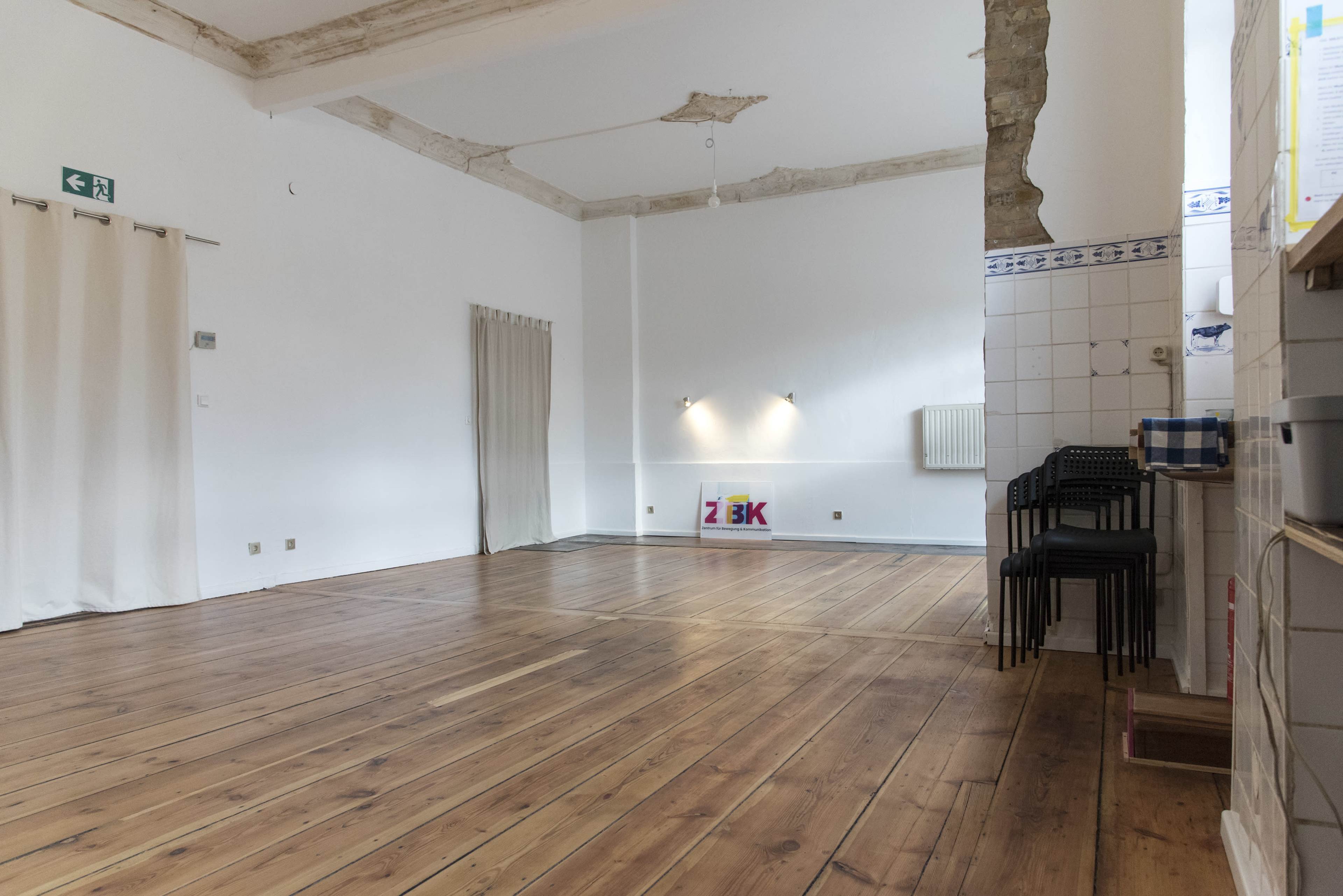 A spacious, empty room with wooden floors, white walls, and minimal furniture against one side.