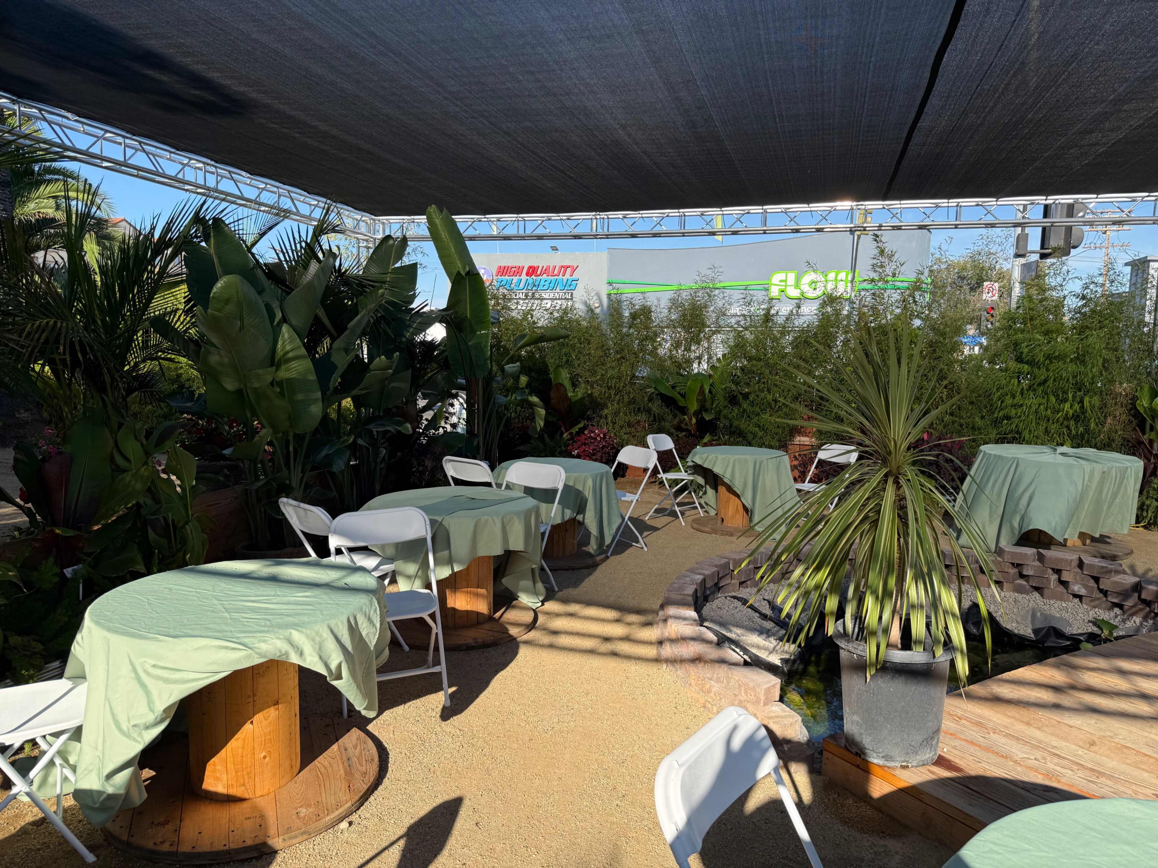 The image shows an outdoor event space with several tables covered in green cloth, surrounded by lush plants and a shaded structure above.