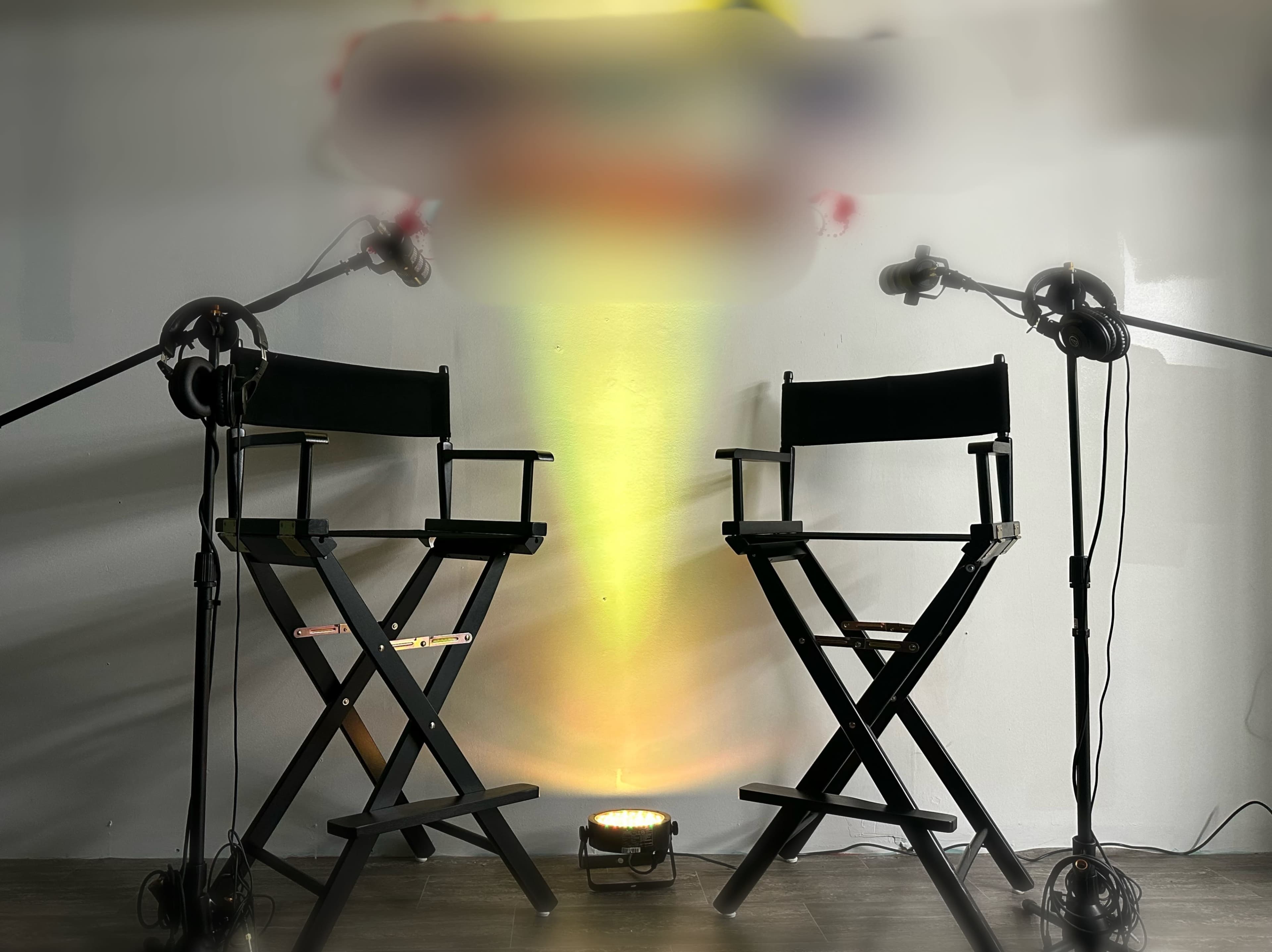 Two director's chairs are positioned facing each other under a spotlight, with microphones set on stands beside them.