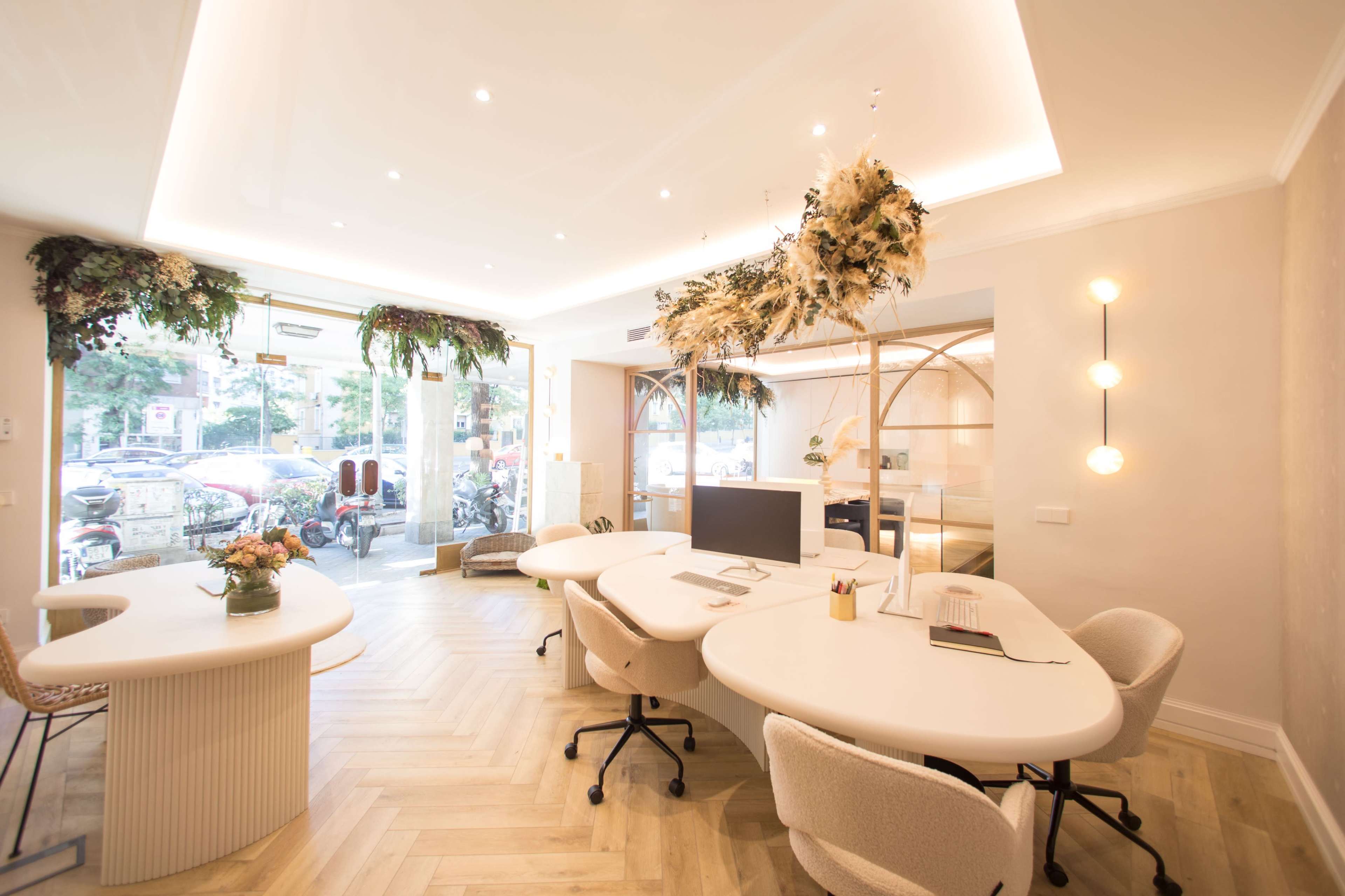 The image shows a modern office space with white furniture, large windows, and decorative plants along the ceiling.