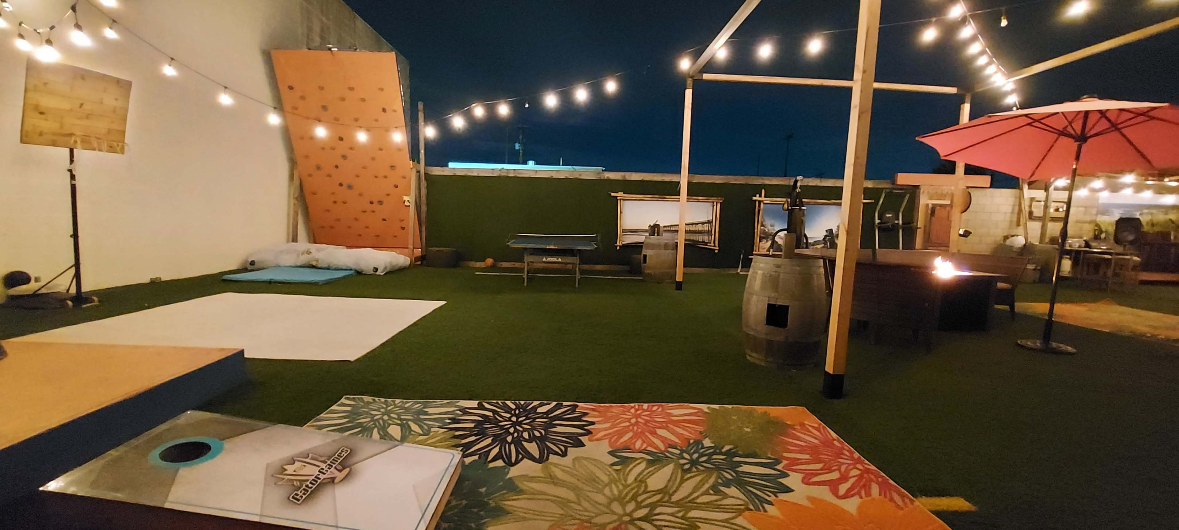 An outdoor recreational area with twinkling lights, a rock climbing wall, seating areas, and various games set up on a grassy surface.