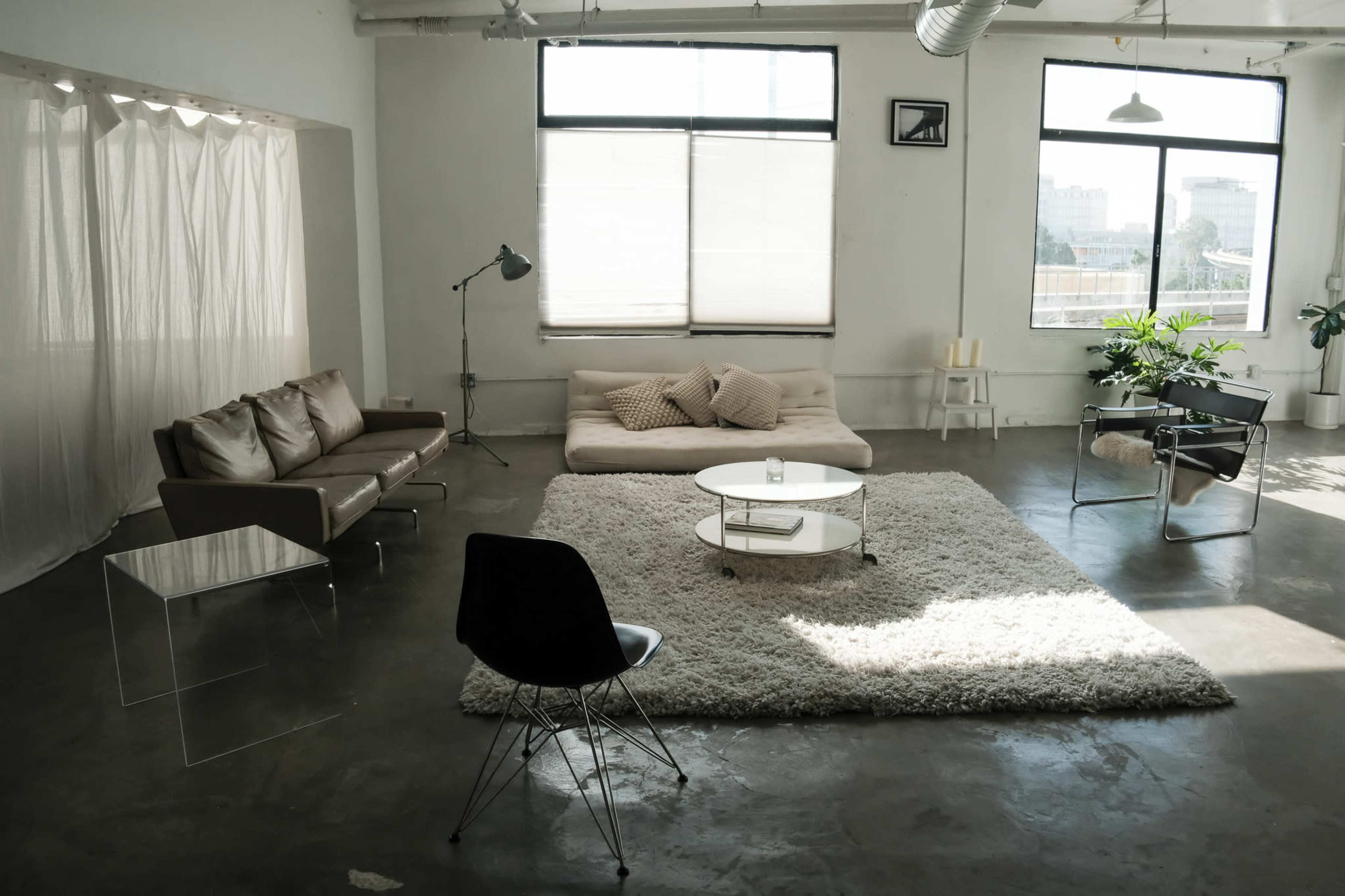 The image shows a modern, minimalist living space featuring a brown sofa, a white rug, a coffee table, and a large window letting in natural light.