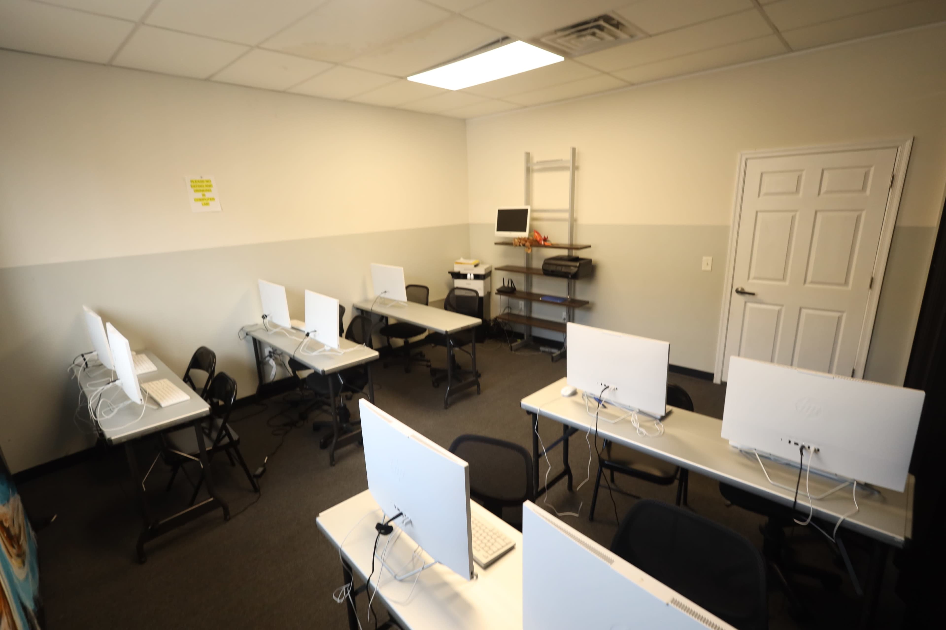 The image shows a small classroom with several empty desks, each equipped with a computer, and a bookshelf against the wall.