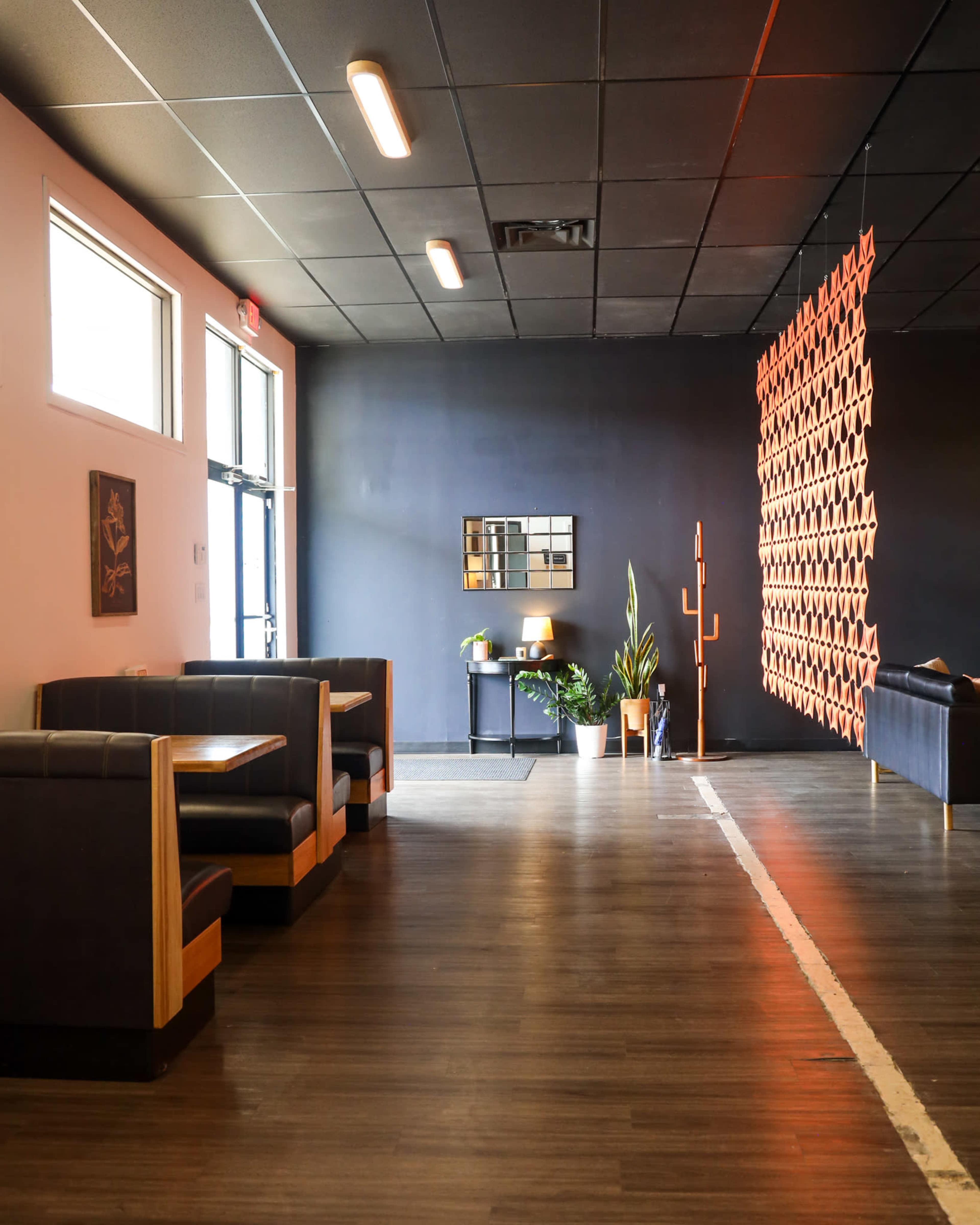 The image shows a modern interior space with seating booths along one wall, a decorative partition, and various plants placed around the room.
