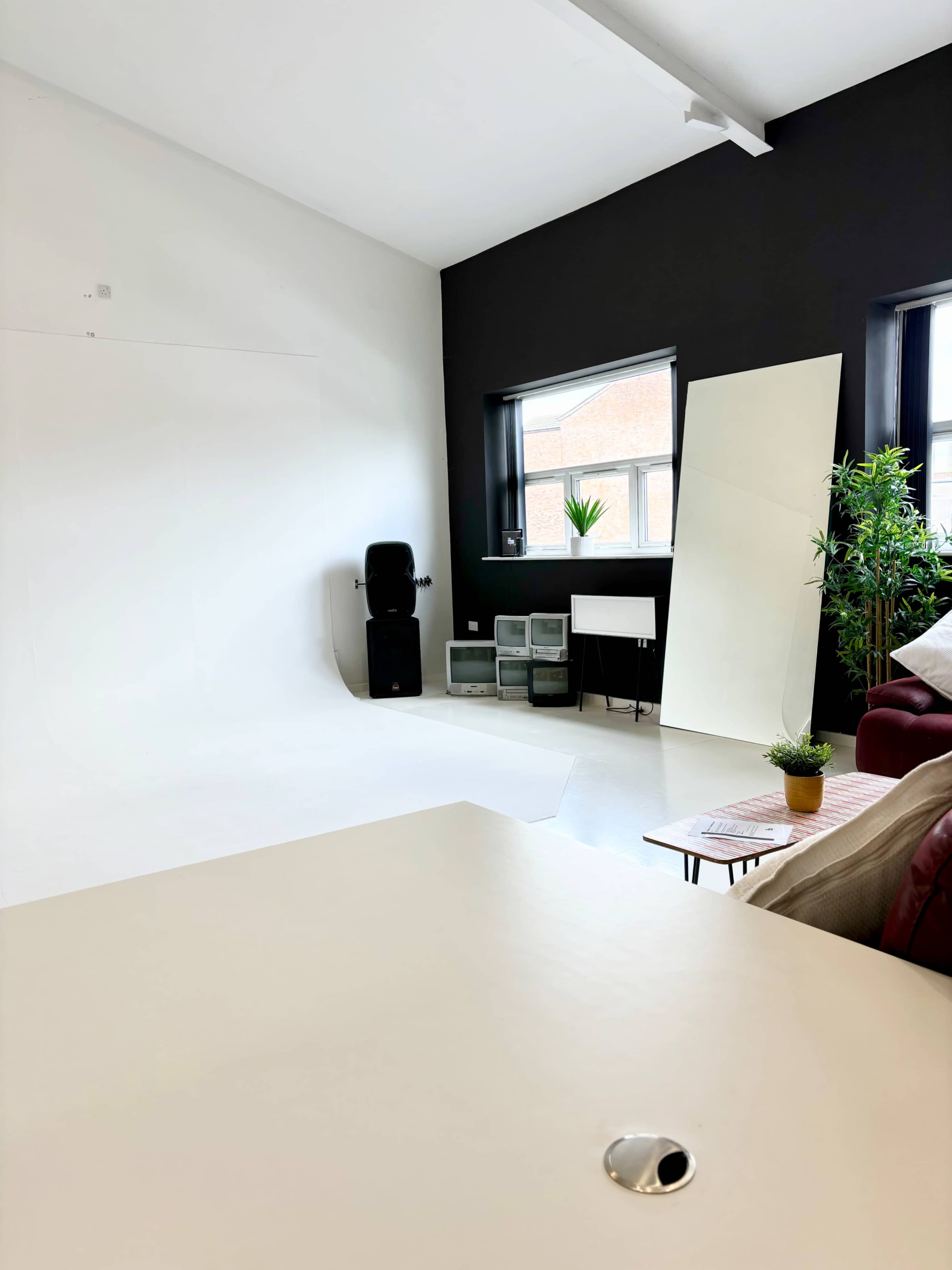 A spacious studio with a white backdrop, a black accent wall, a potted plant, a seating area, and storage boxes.